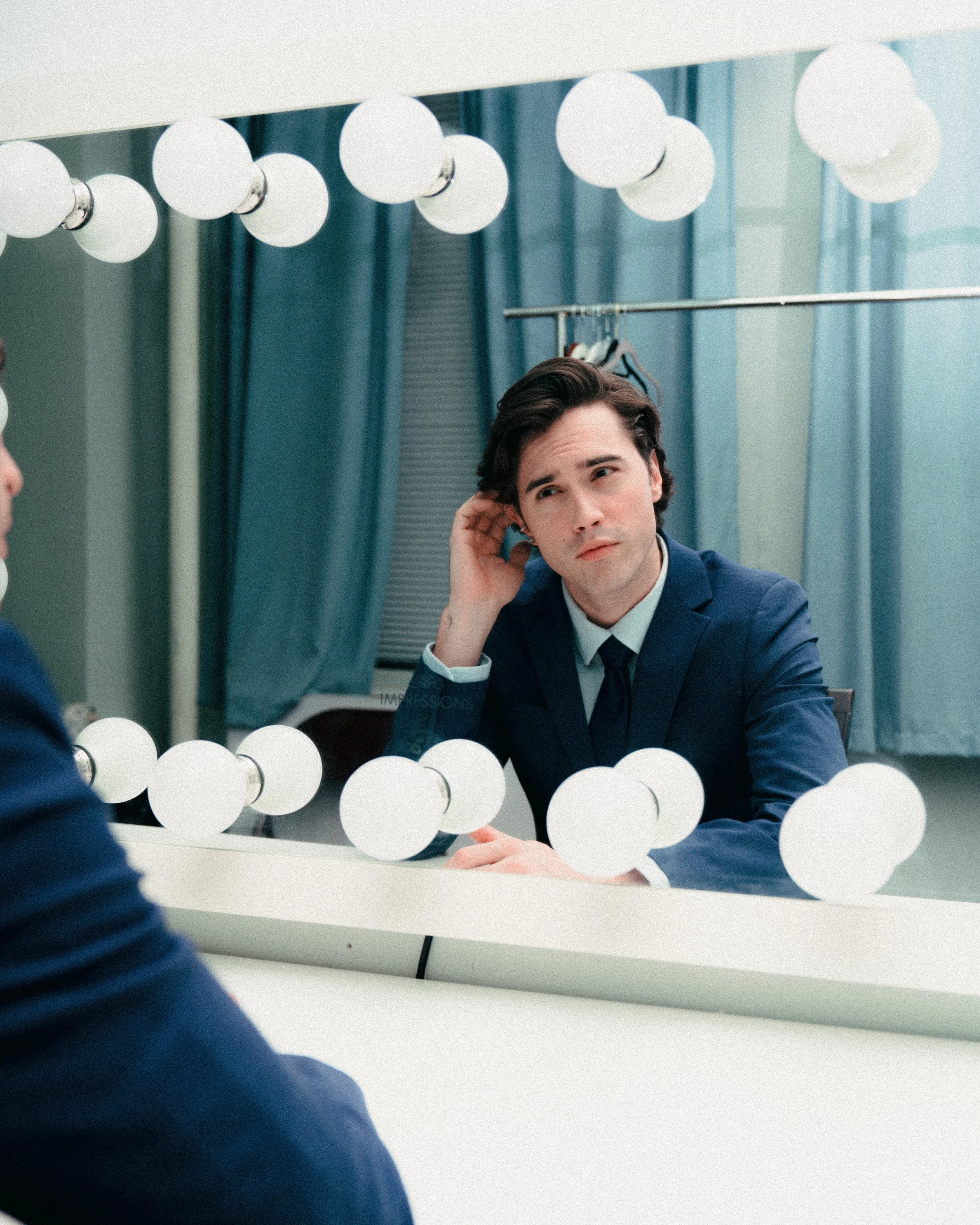 Young man in a blue suit looking at himself in a mirror with bright bulbs around it, touching his ear thoughtfully.