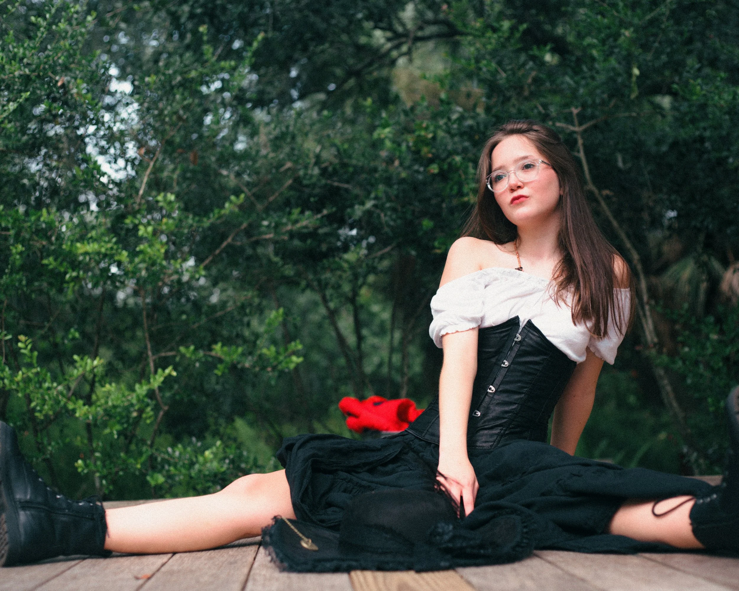 A young woman with glasses and long brown hair sitting on a wooden deck outdoors surrounded by green bushes, dressed in a black corset and black skirt, with a white off-shoulder top.