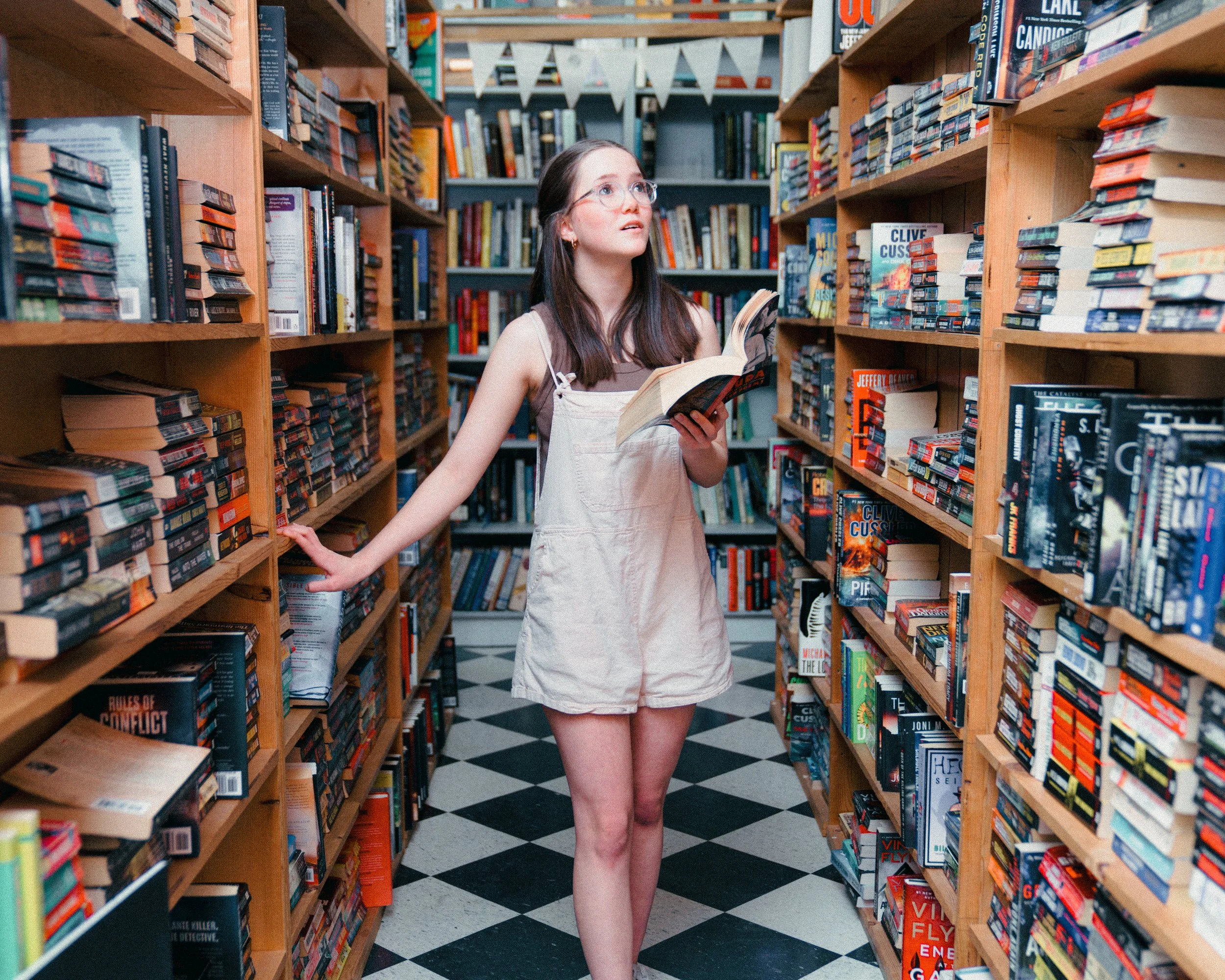 A young woman with glasses and long brown hair browsing books in a bookstore aisle, wearing a sleeveless top and a light-colored overall dress, with bookshelves filled with books on either side.