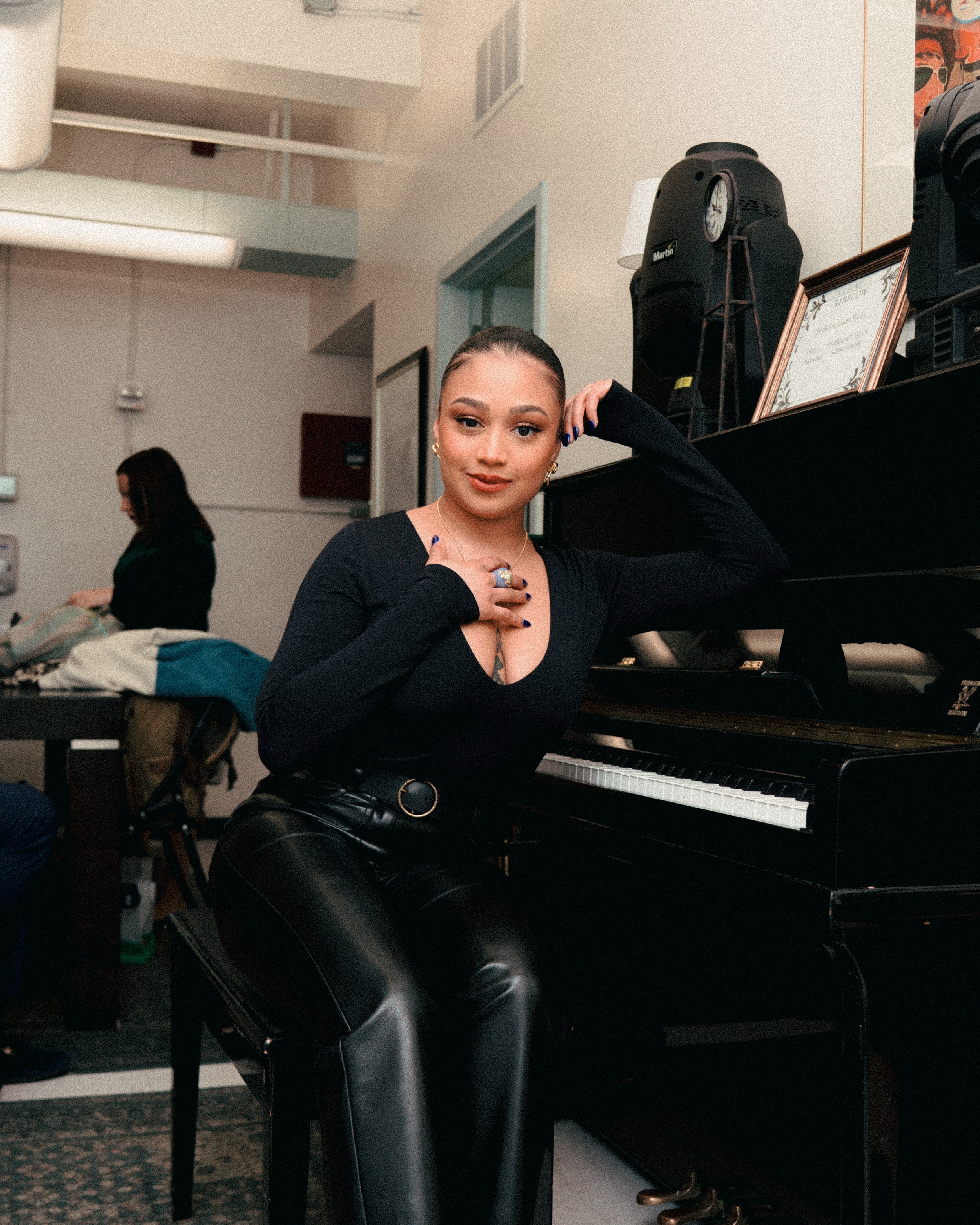 Young woman with makeup and jewelry sitting at a black upright piano, resting her arm on it and looking at the camera, in a room with another woman in the background and a framed certificate on the piano.