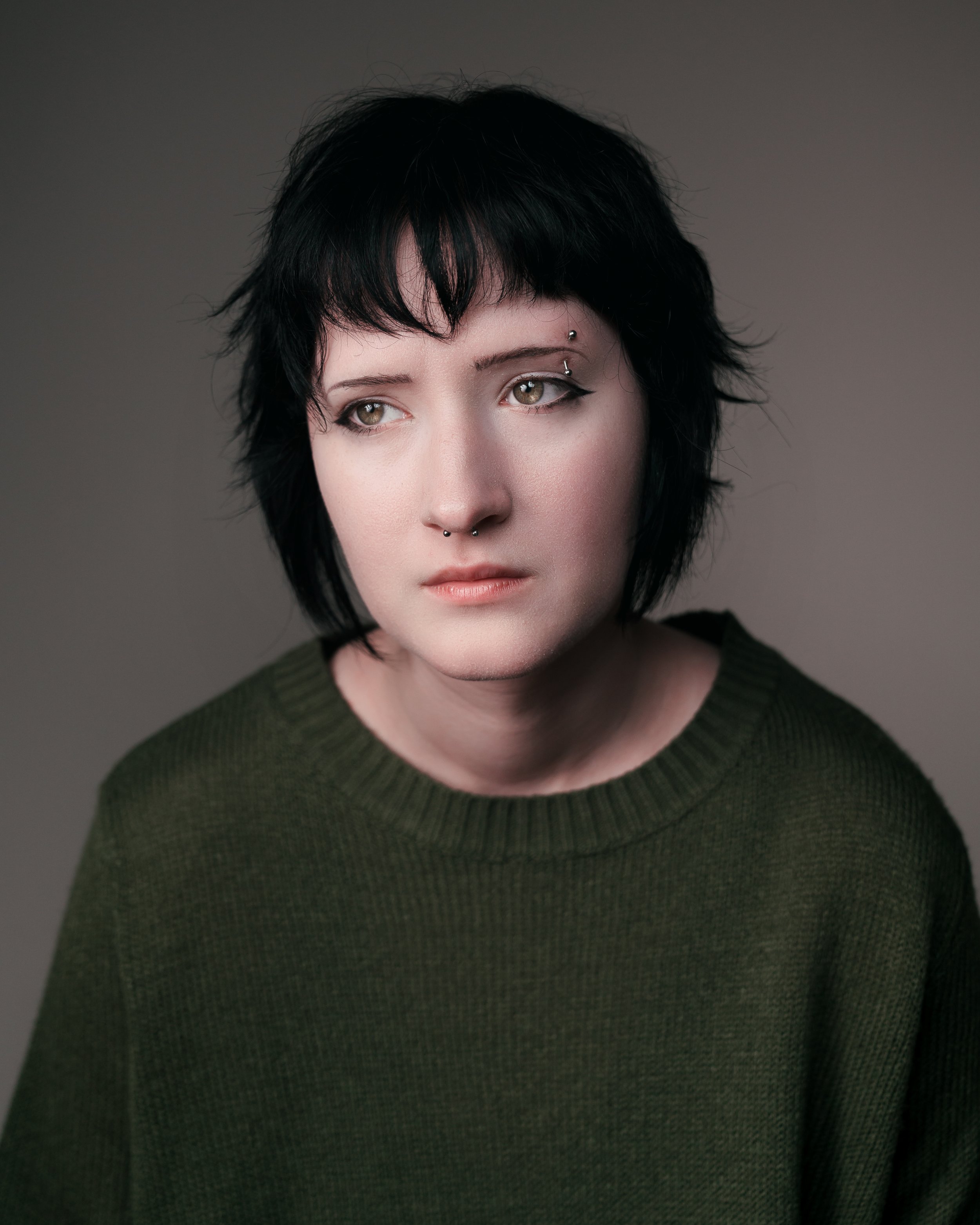 Portrait of a young woman with short black hair, wearing a green sweater, looking to the side with a neutral expression.
