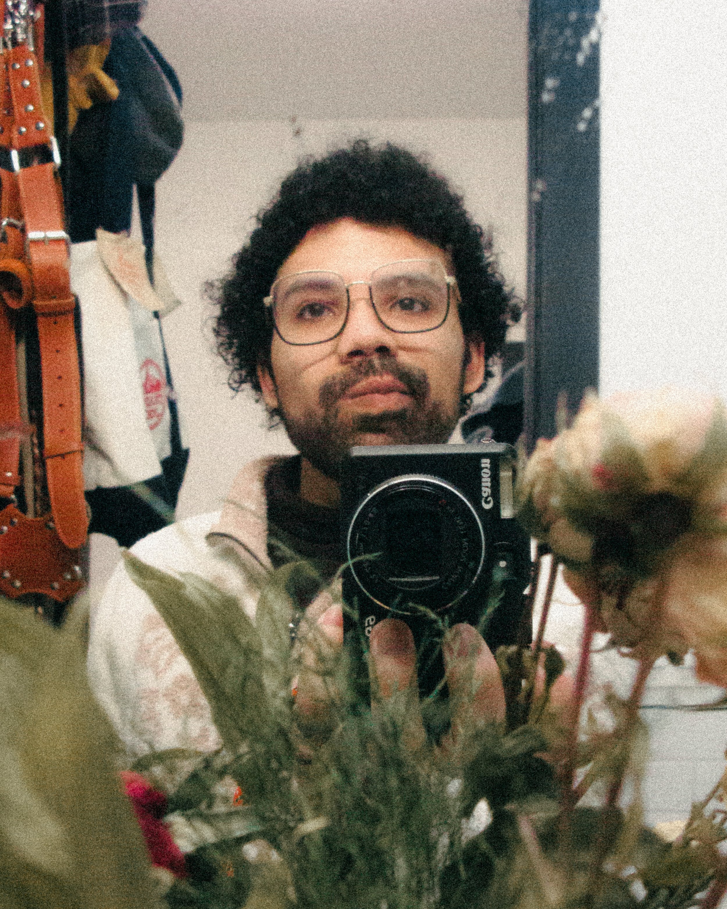 A person with glasses and curly hair takes a selfie in a mirror, with dried flowers and a coat hook in the foreground.
