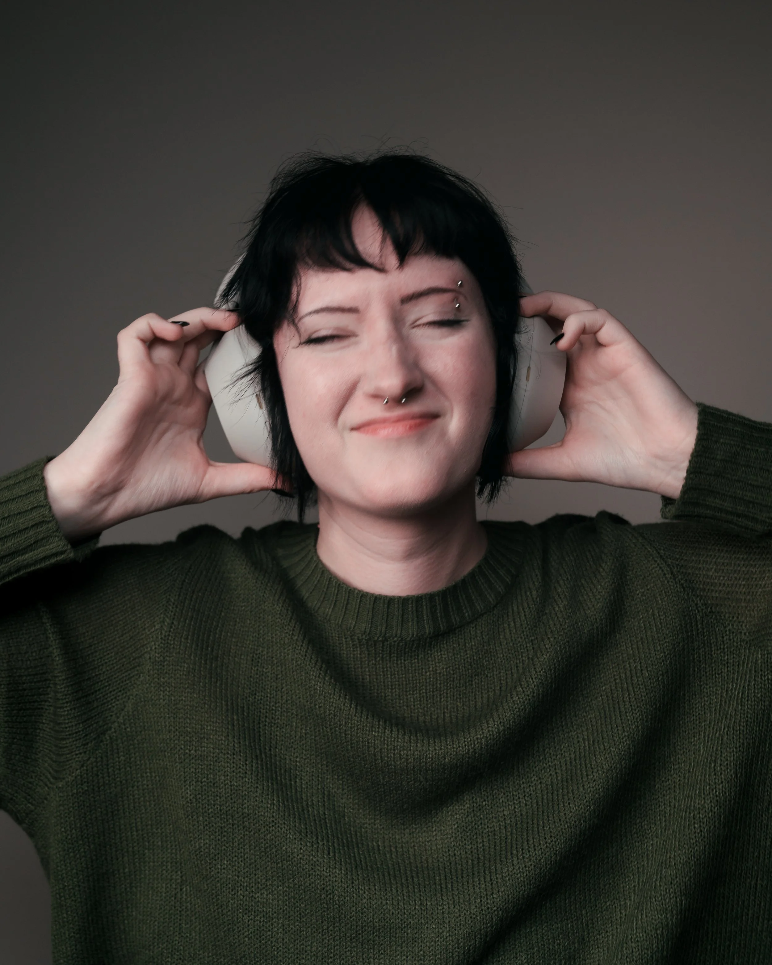 Woman with short black hair wearing a green sweater, with a facial expression of enjoyment while listening to music through white headphones.