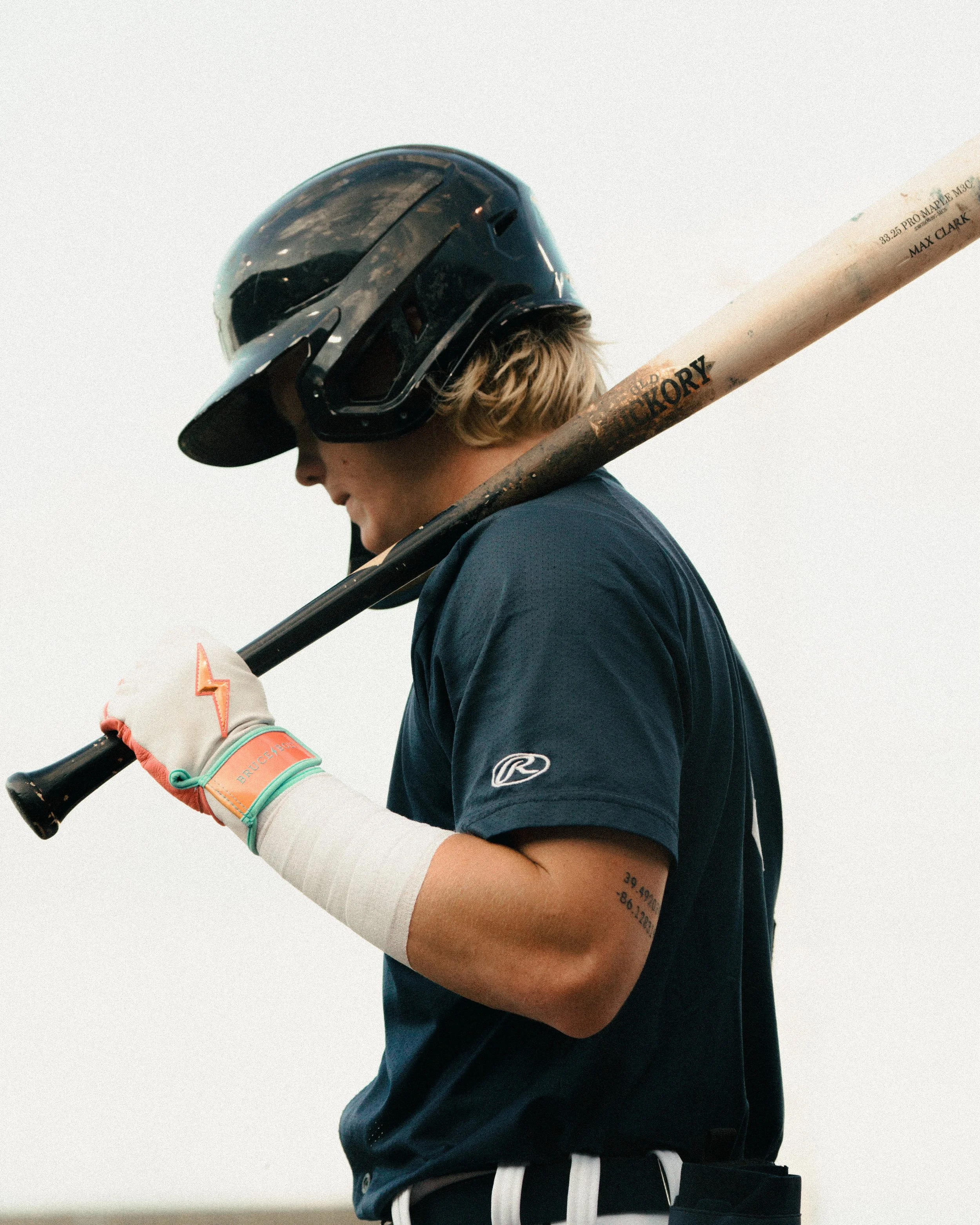 A young baseball player wearing a helmet, holding a bat over his shoulder, standing outdoors against a gray sky.