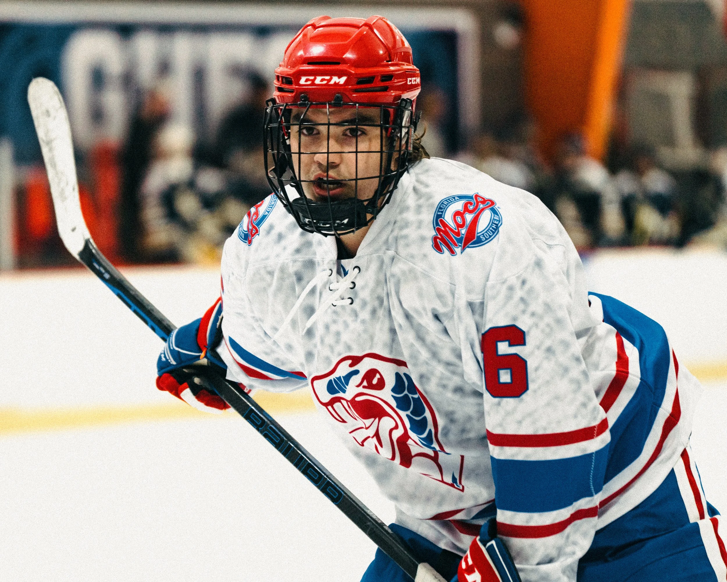 An ice hockey player wearing a white jersey with red and blue accents, a red helmet, and blue gloves, holding a hockey stick on the ice.