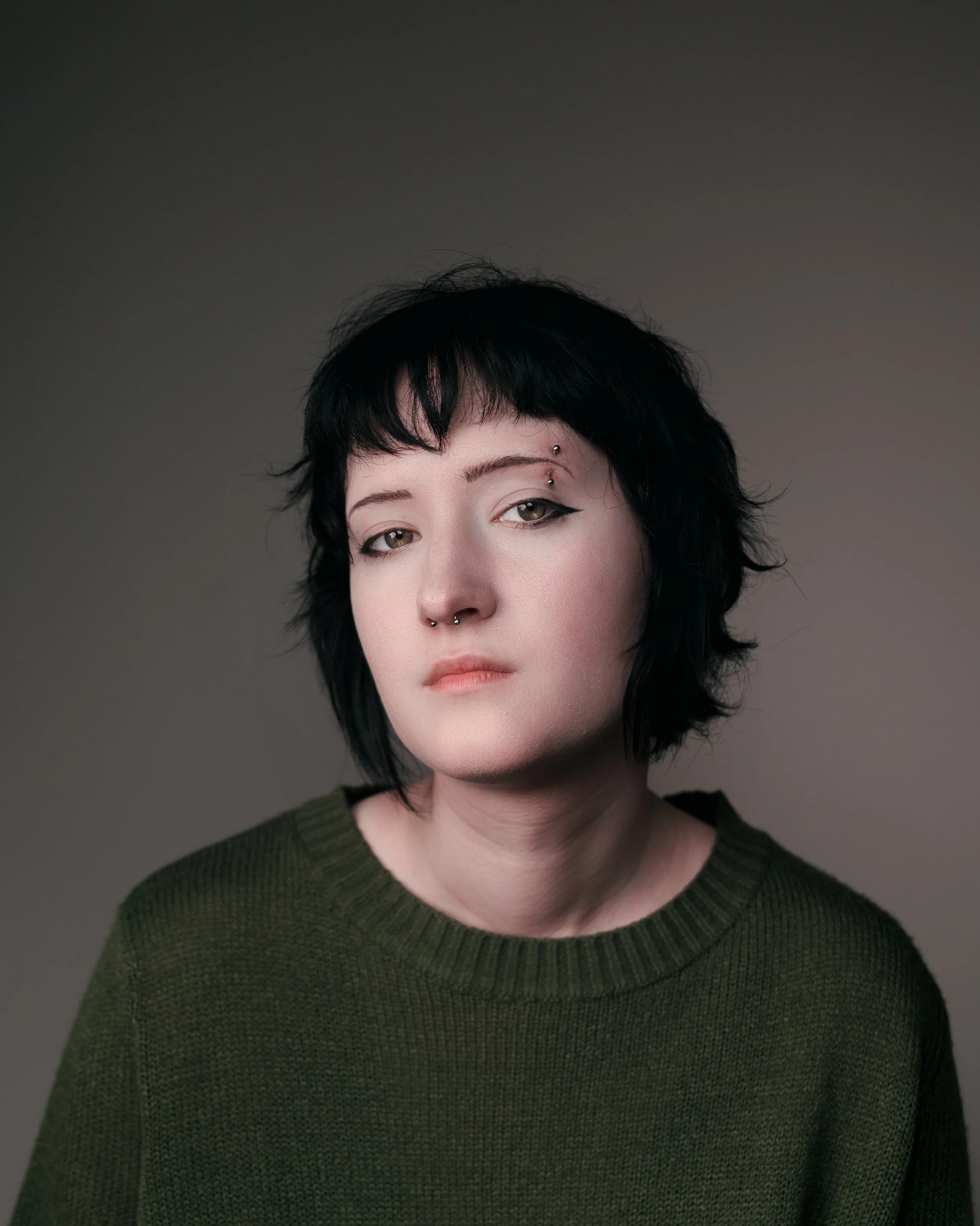 Portrait of a young woman with short black hair and piercings, wearing a green sweater against a gray background.