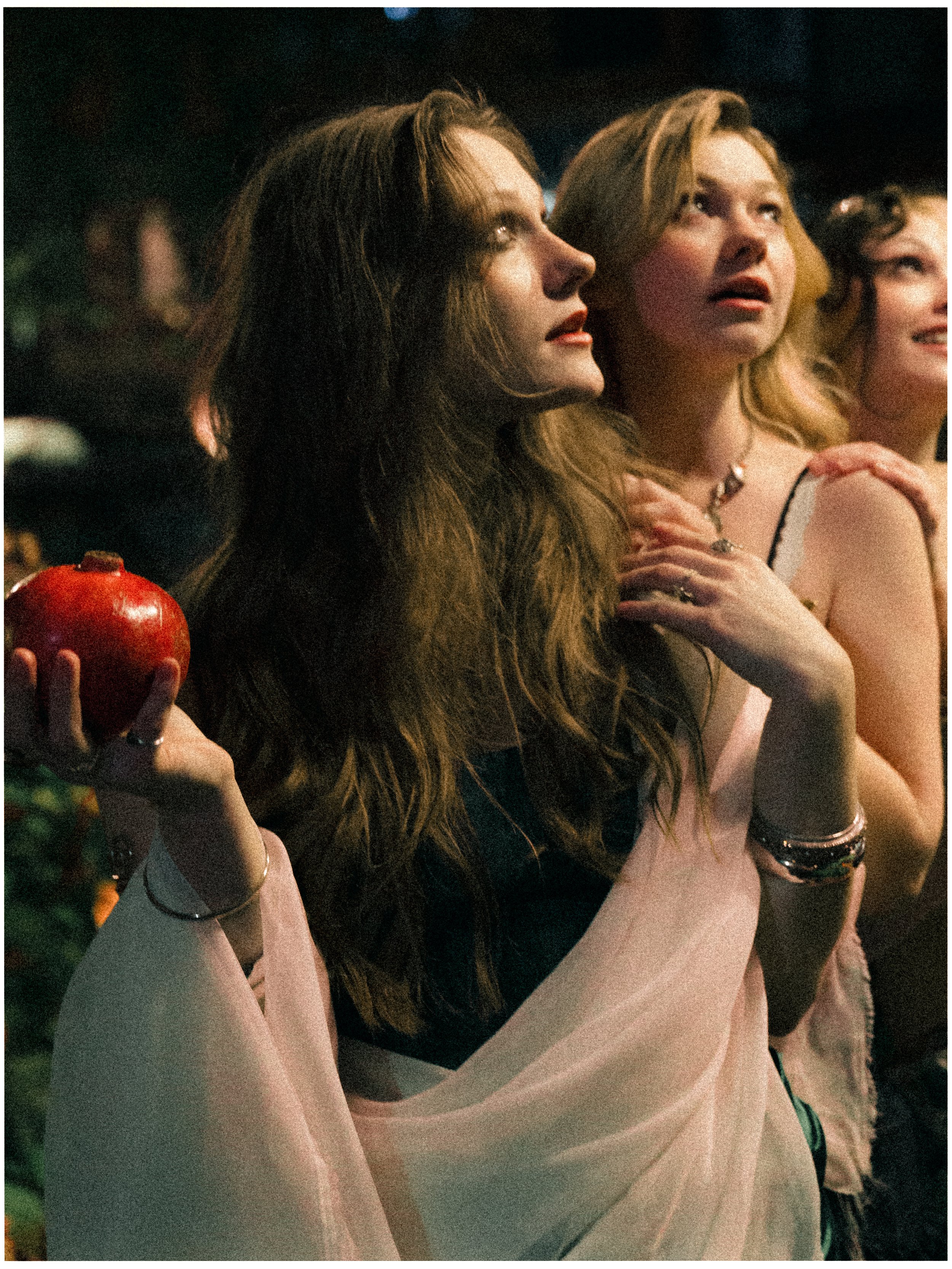 Three women sitting closely together at a social event, with one woman in the foreground holding a red apple and wearing a light pink shawl, while the other two women are looking upward with expressions of wonder or surprise.