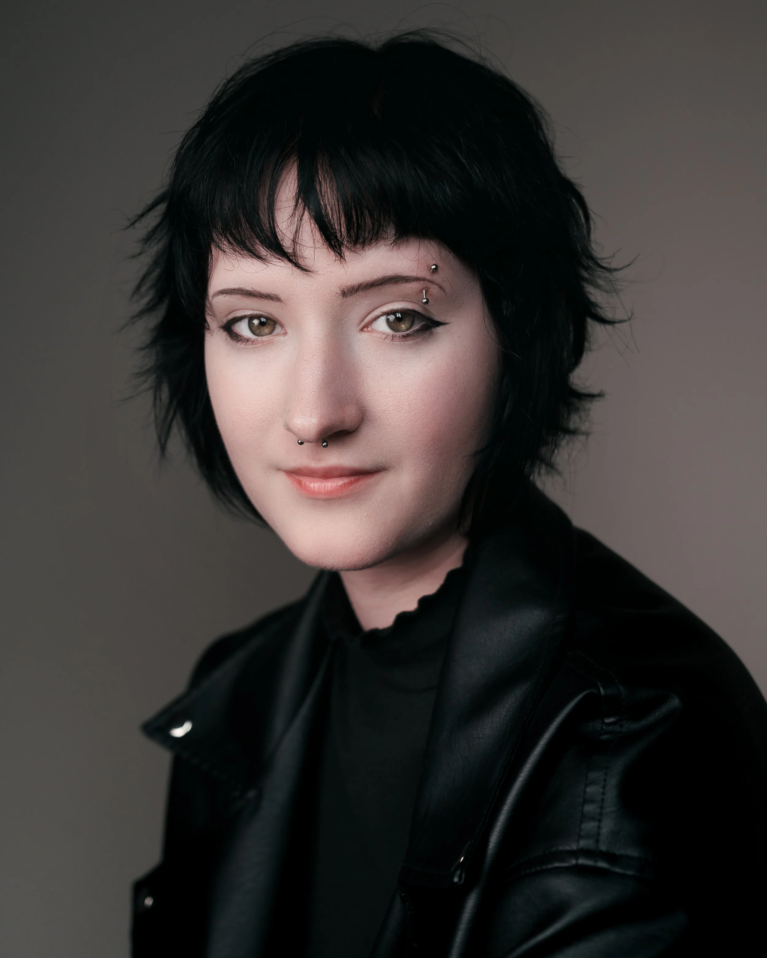 Portrait of a young woman with short black hair, multiple facial piercings, wearing a black leather jacket and black top, against a plain gray background.