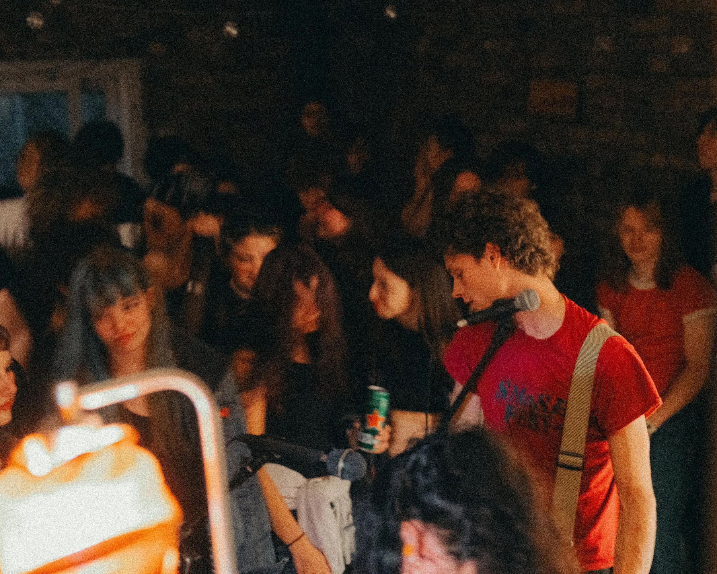 A person with curly hair wearing a red T-shirt with a beige strap over their shoulder, holding a guitar and a microphone, singing or speaking to a crowd in a dimly lit indoor venue.