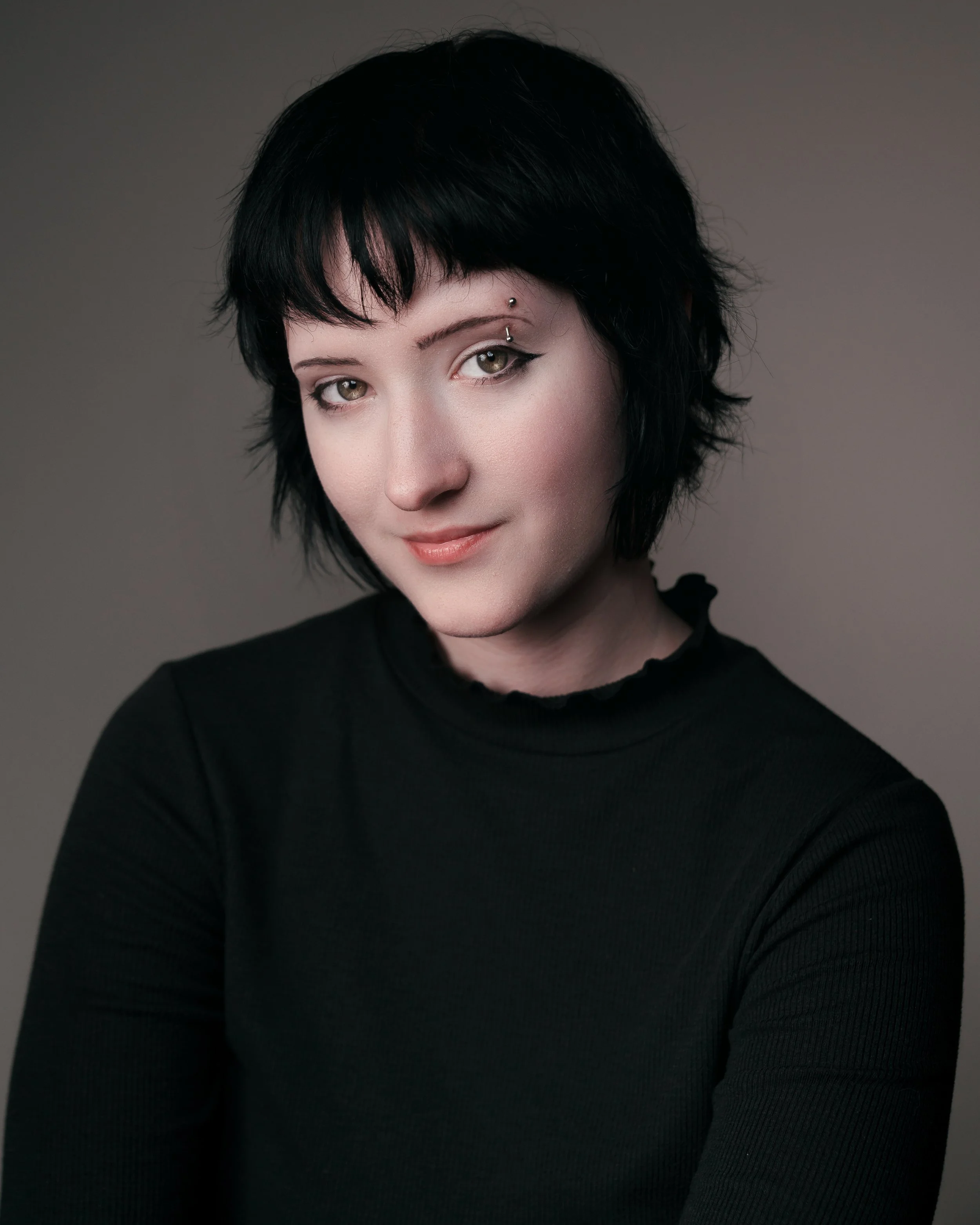 Portrait of a young woman with short black hair, wearing a black top, looking at the camera with a slight smile against a plain gray background.
