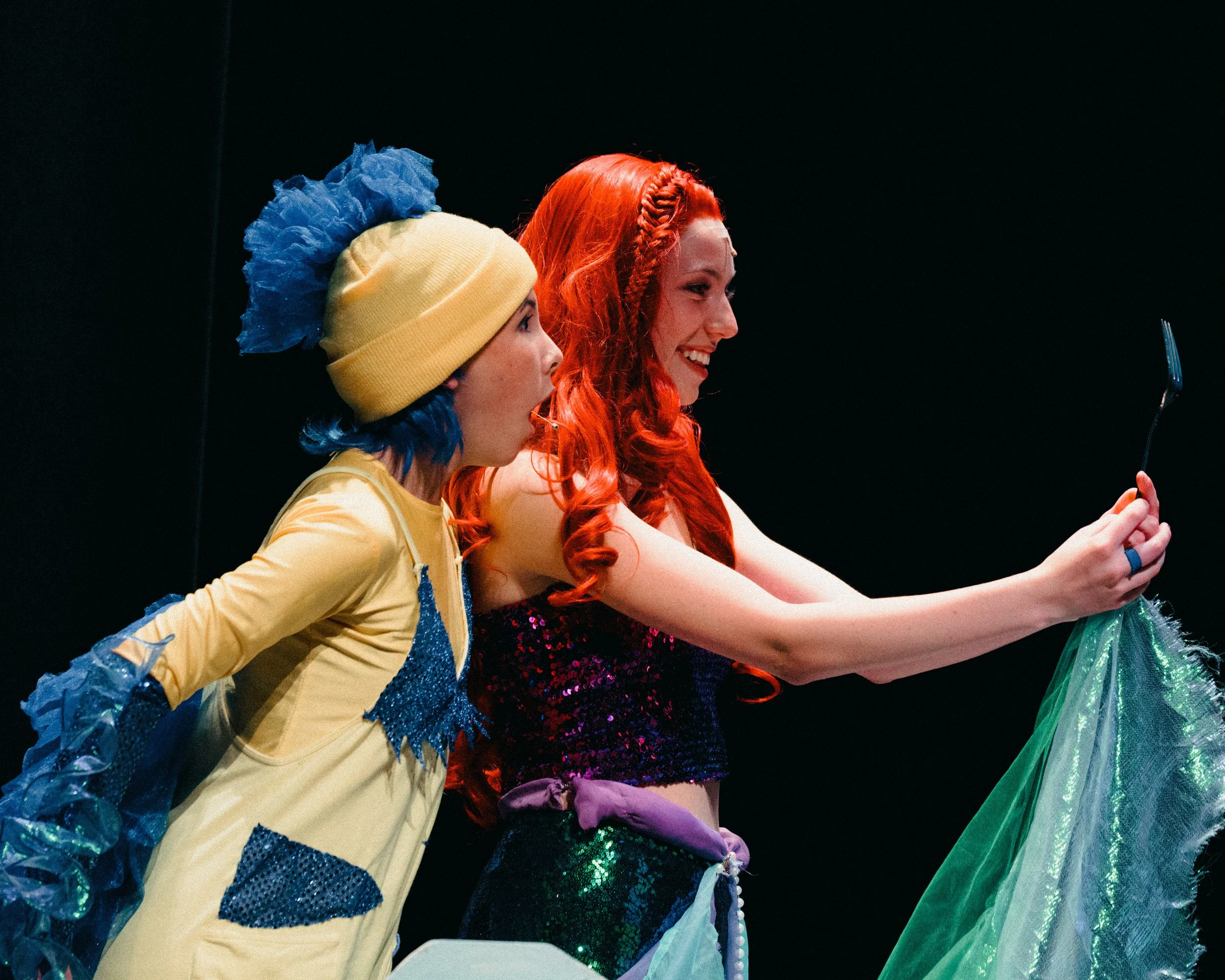 Two women in colorful costumes taking a selfie on stage, with a black background.