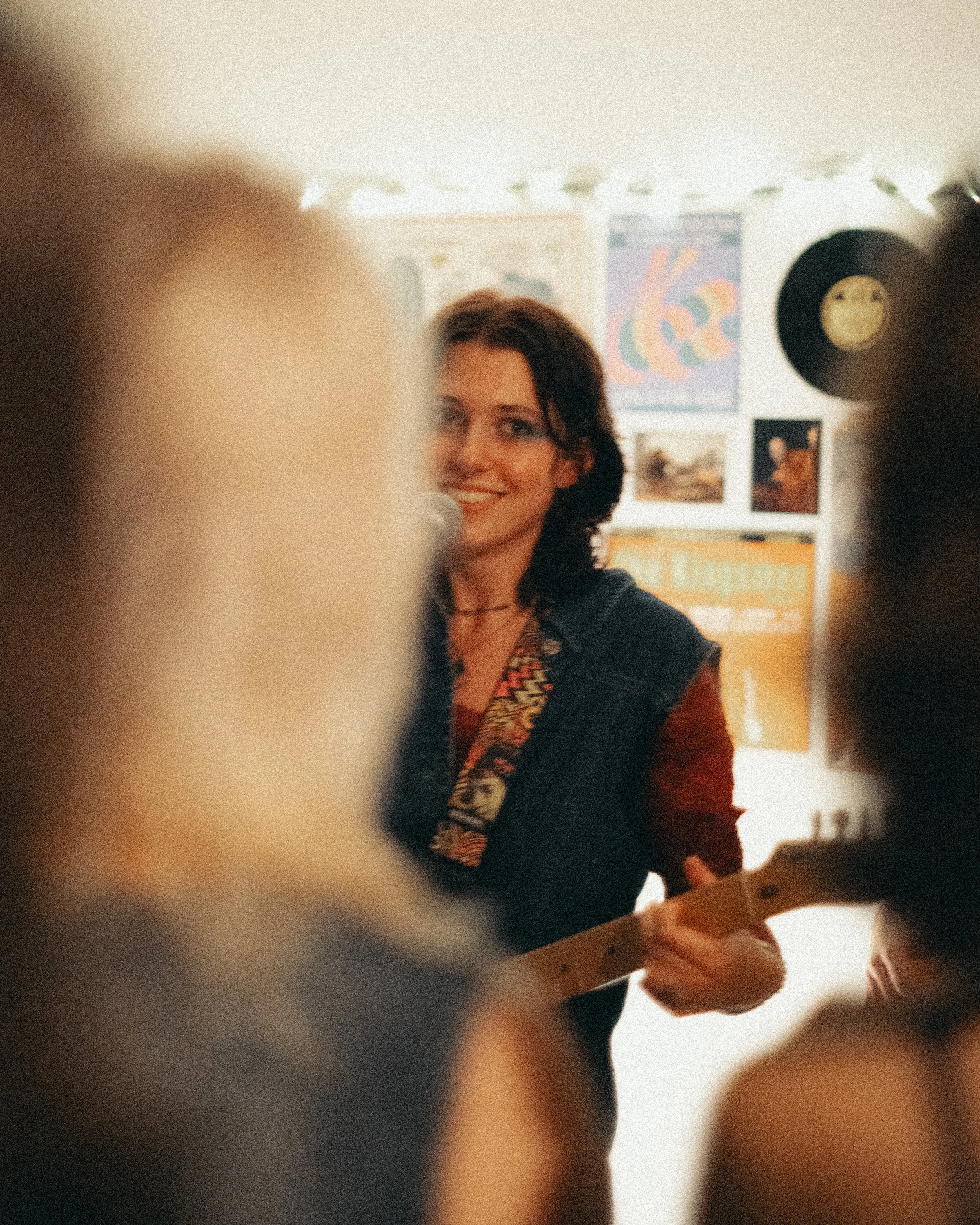 A woman with dark, curly hair smiling while playing a guitar in a room decorated with colorful artwork and records on the wall.
