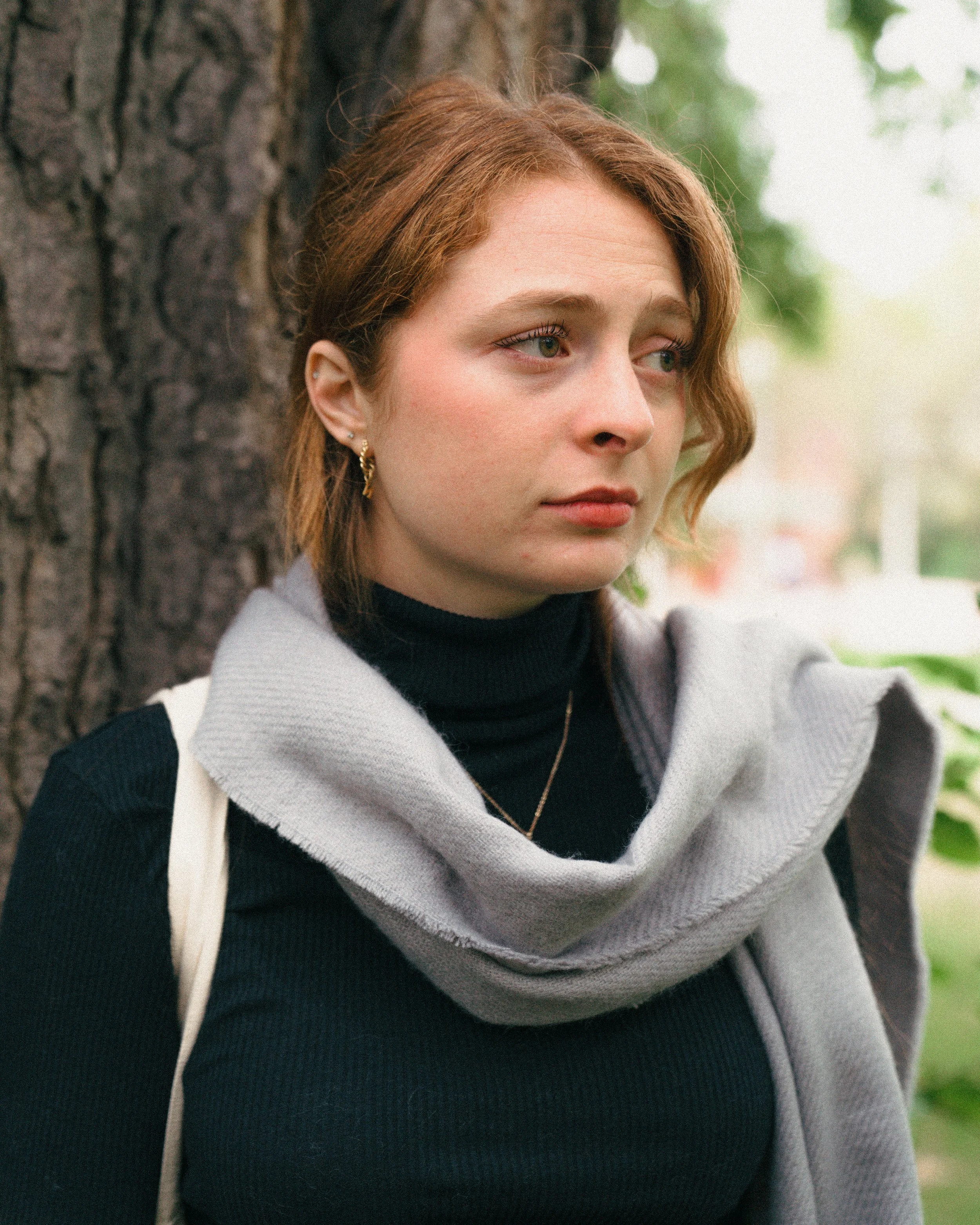 A young woman with red hair and earrings standing outdoors next to a tree, wearing a black turtleneck and a gray scarf, looking contemplative.