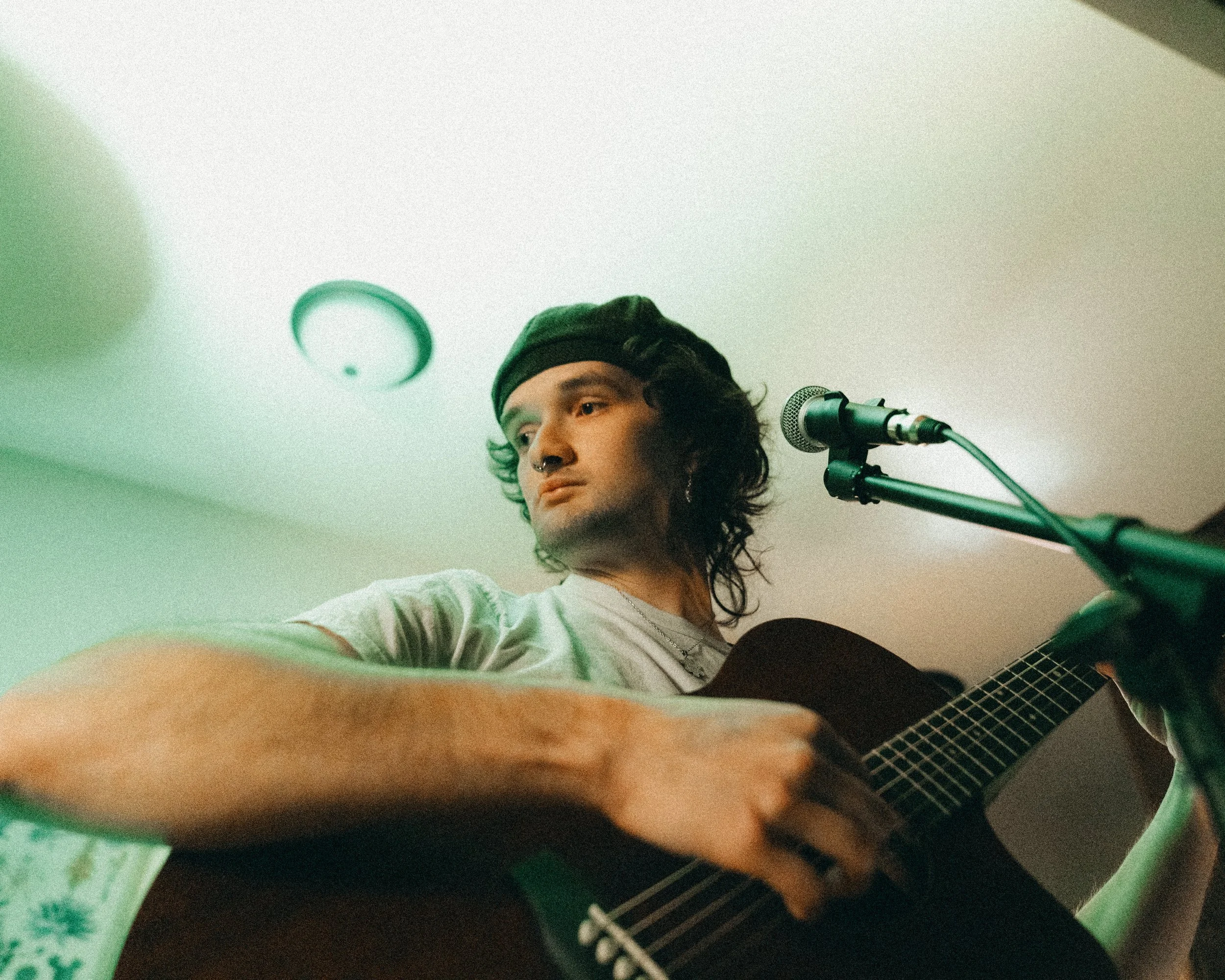 A young man with curly hair, wearing a black beanie and a short-sleeved shirt, playing an acoustic guitar in front of a microphone with a neutral expression.