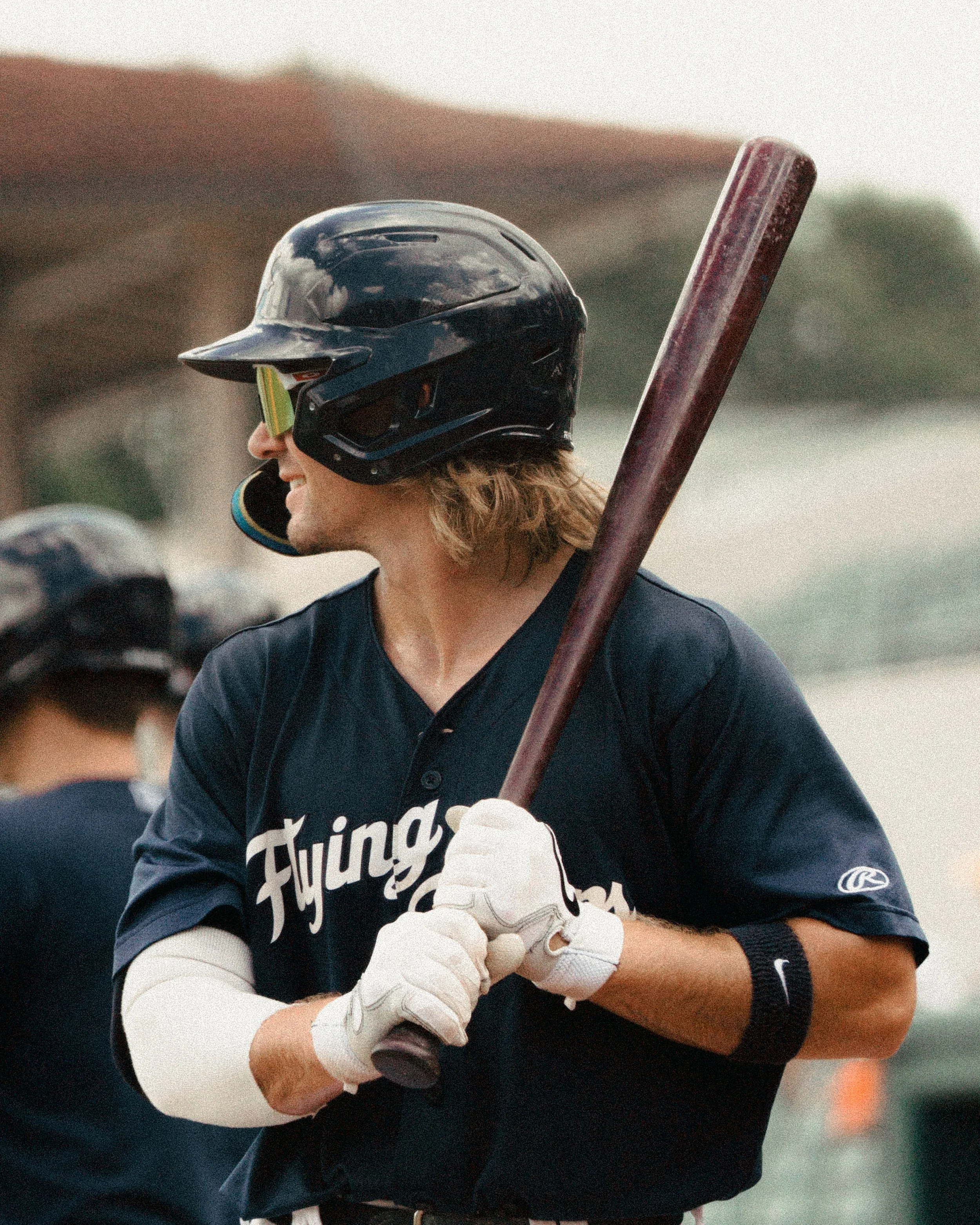 A baseball player with long hair wearing a navy blue uniform and a black helmet holding a bat over his shoulder, standing outdoors.