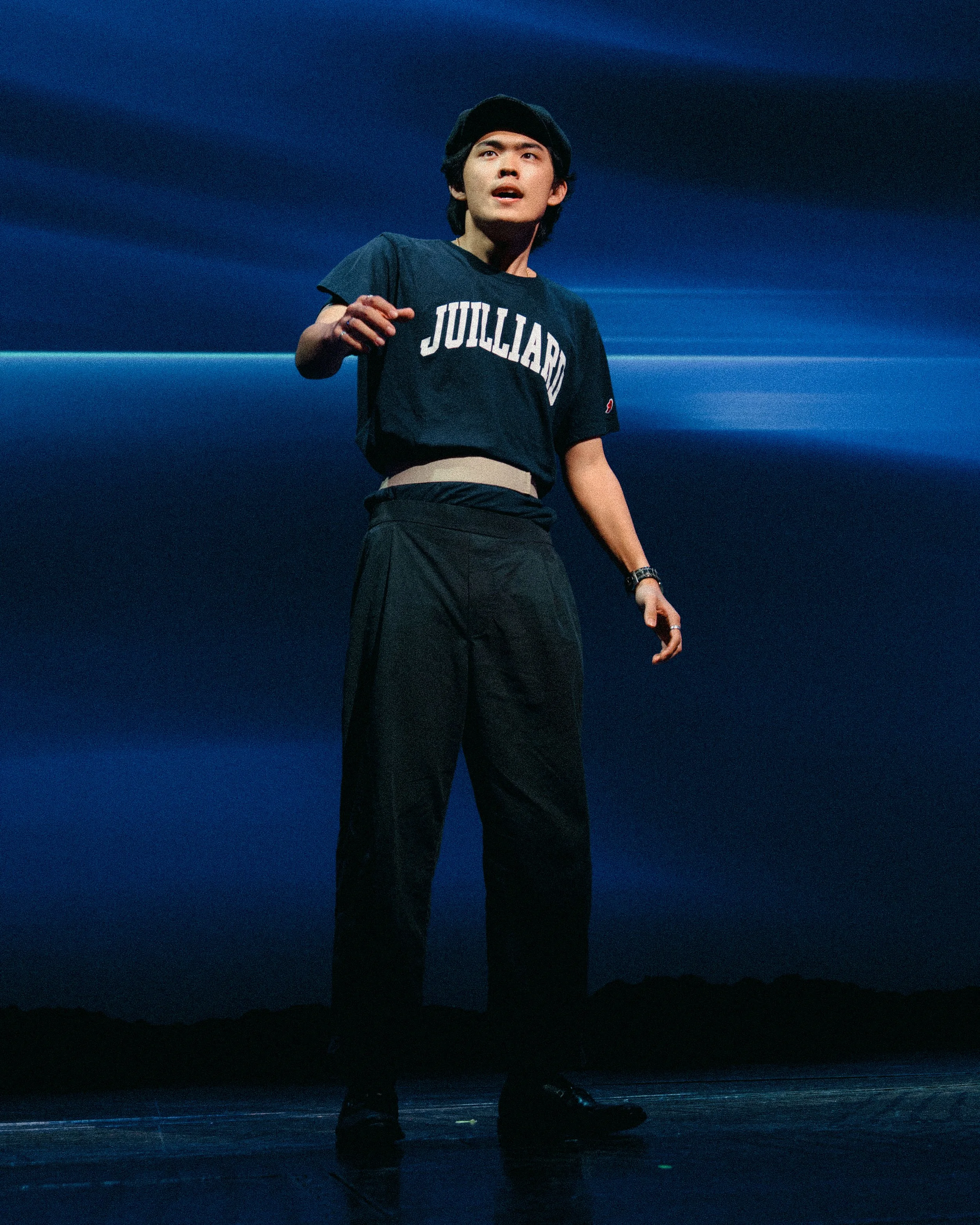 A young man with dark hair, wearing a black cap, a black Julllard T-shirt, black pants, and black shoes, standing on stage against a blue background, speaking or performing.