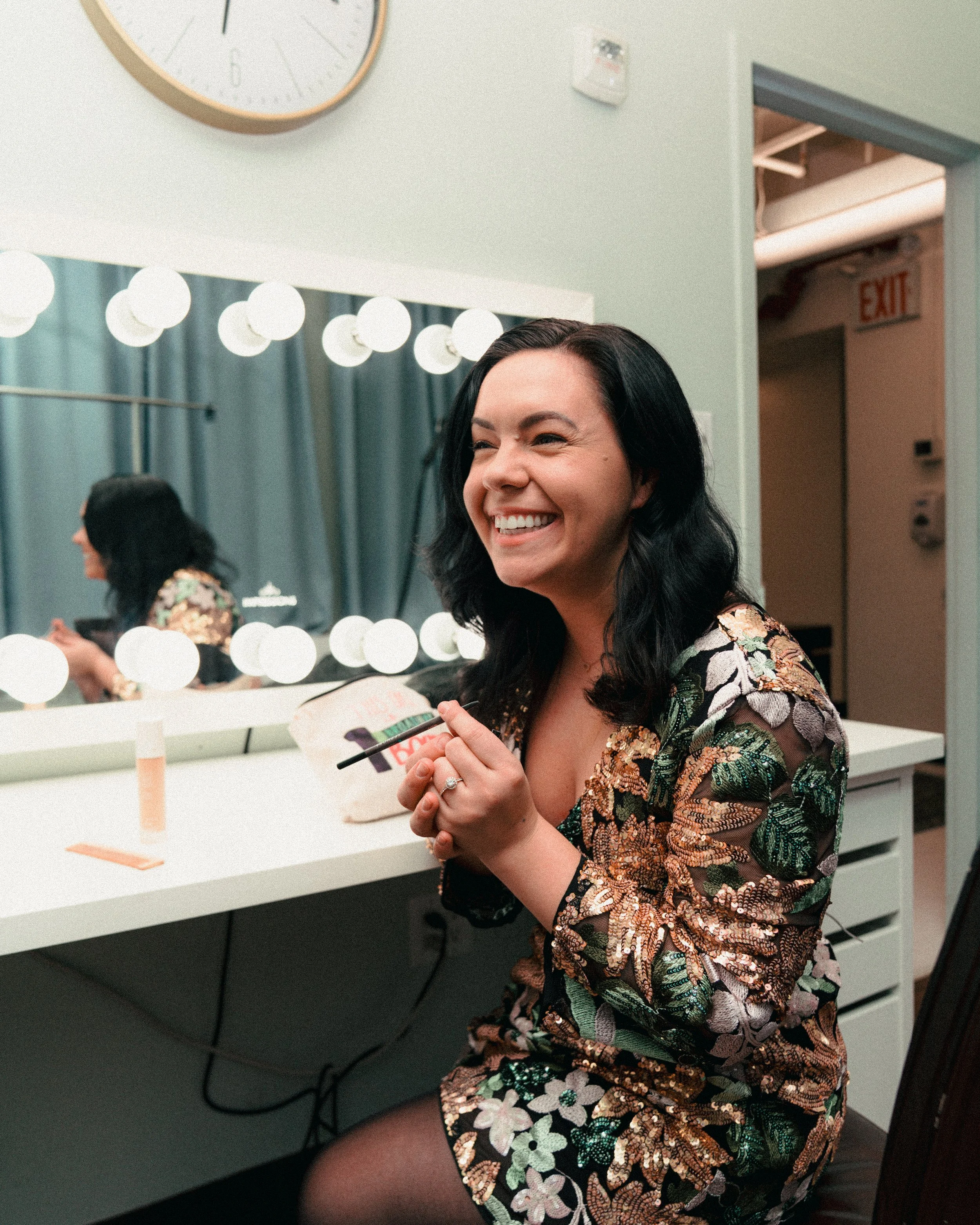 A woman with black hair smiling and holding a small makeup brush sitting in front of a mirror with bright lights in a dressing room or makeup room.