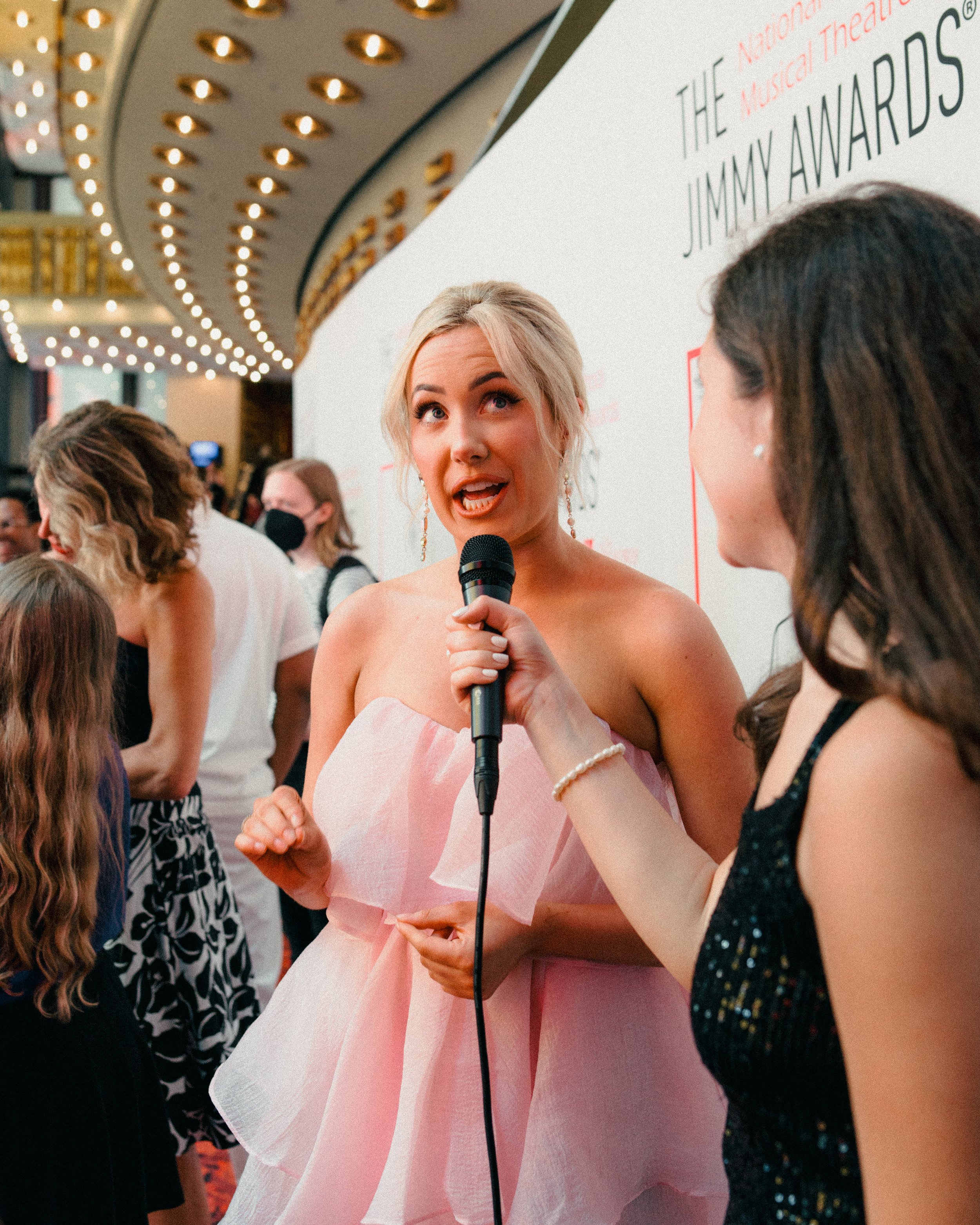 A woman in a strapless pink dress being interviewed with a microphone at an event, with others in line and a decorative marquee in the background.
