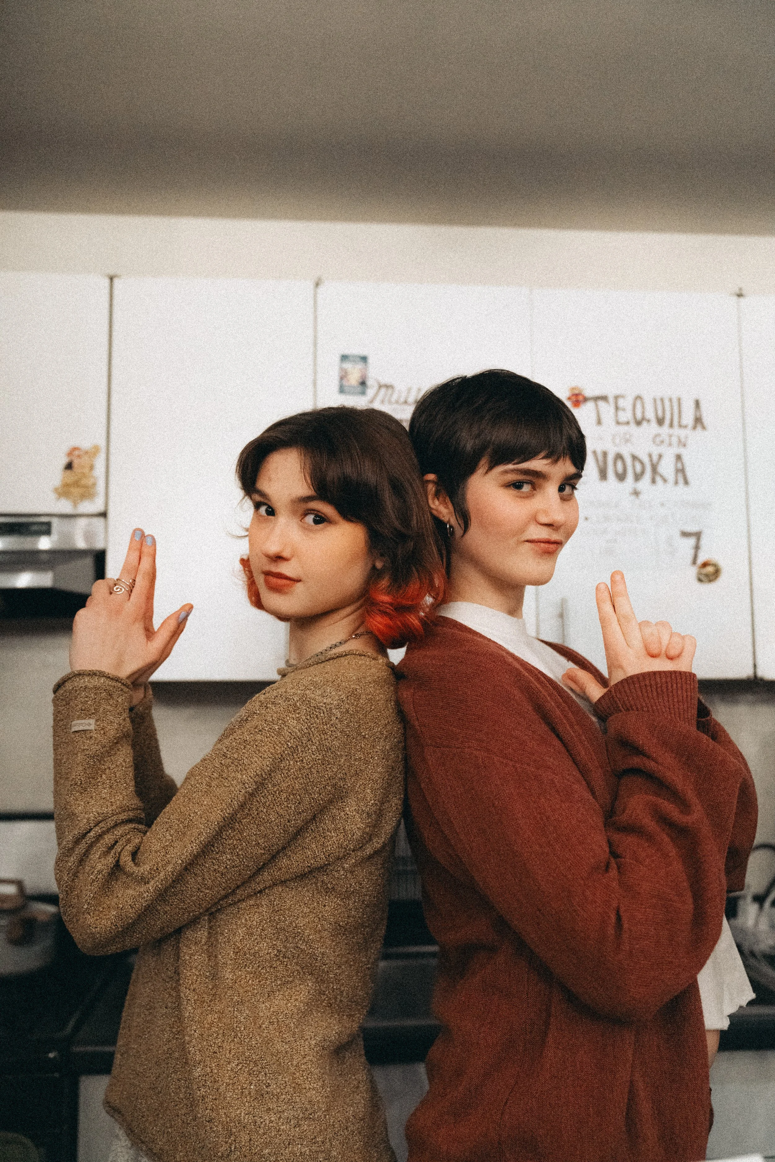 Two young women standing back-to-back in a kitchen, facing the camera, each making a gesture with their hands. One has short dark hair and wears a red sweater, the other has brown hair with red tips and wears a tan sweater.