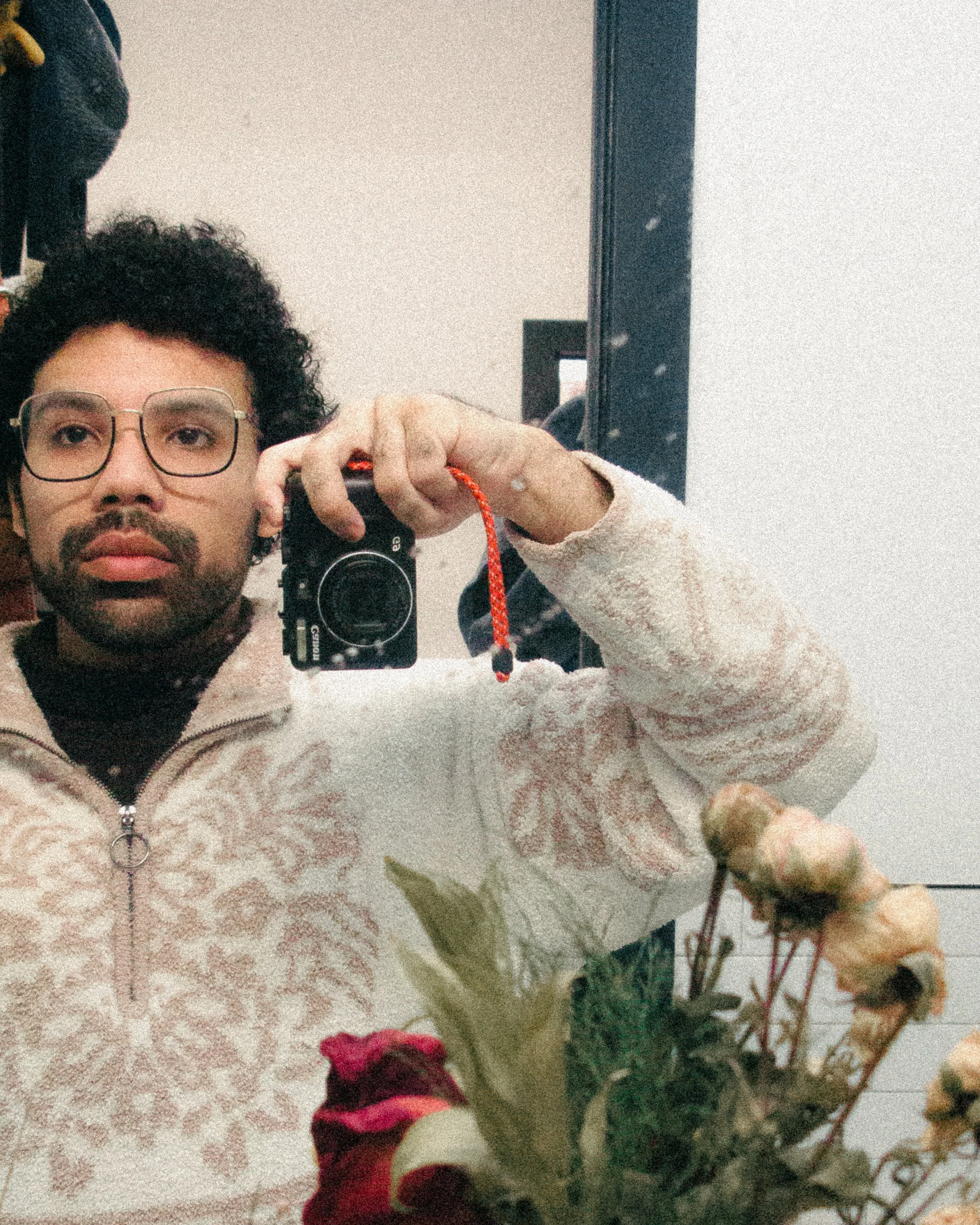 A man with curly hair and glasses taking a mirror selfie with a camera, wearing a beige fleece jacket with a floral pattern, holding a bouquet of flowers.