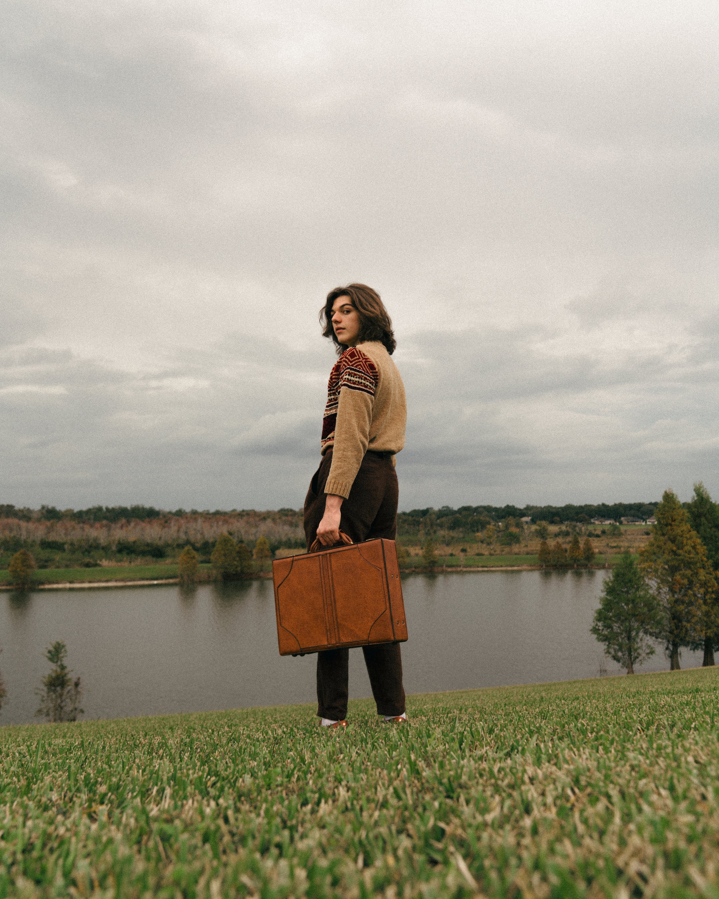 A woman standing on a grassy hill near a body of water, holding a leather suitcase, with trees and overcast sky in the background.