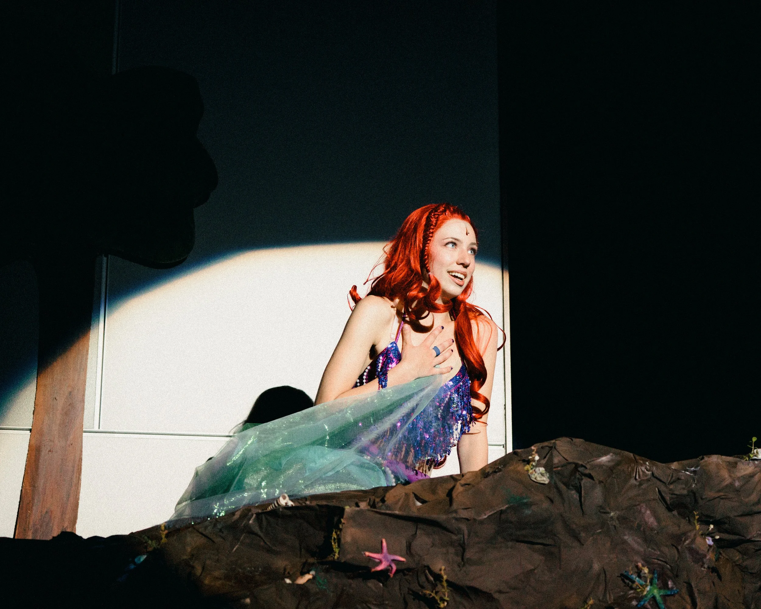 Young woman with red hair dressed as Ariel from The Little Mermaid, on stage under spotlight, with painted rocks and small figurines in foreground.