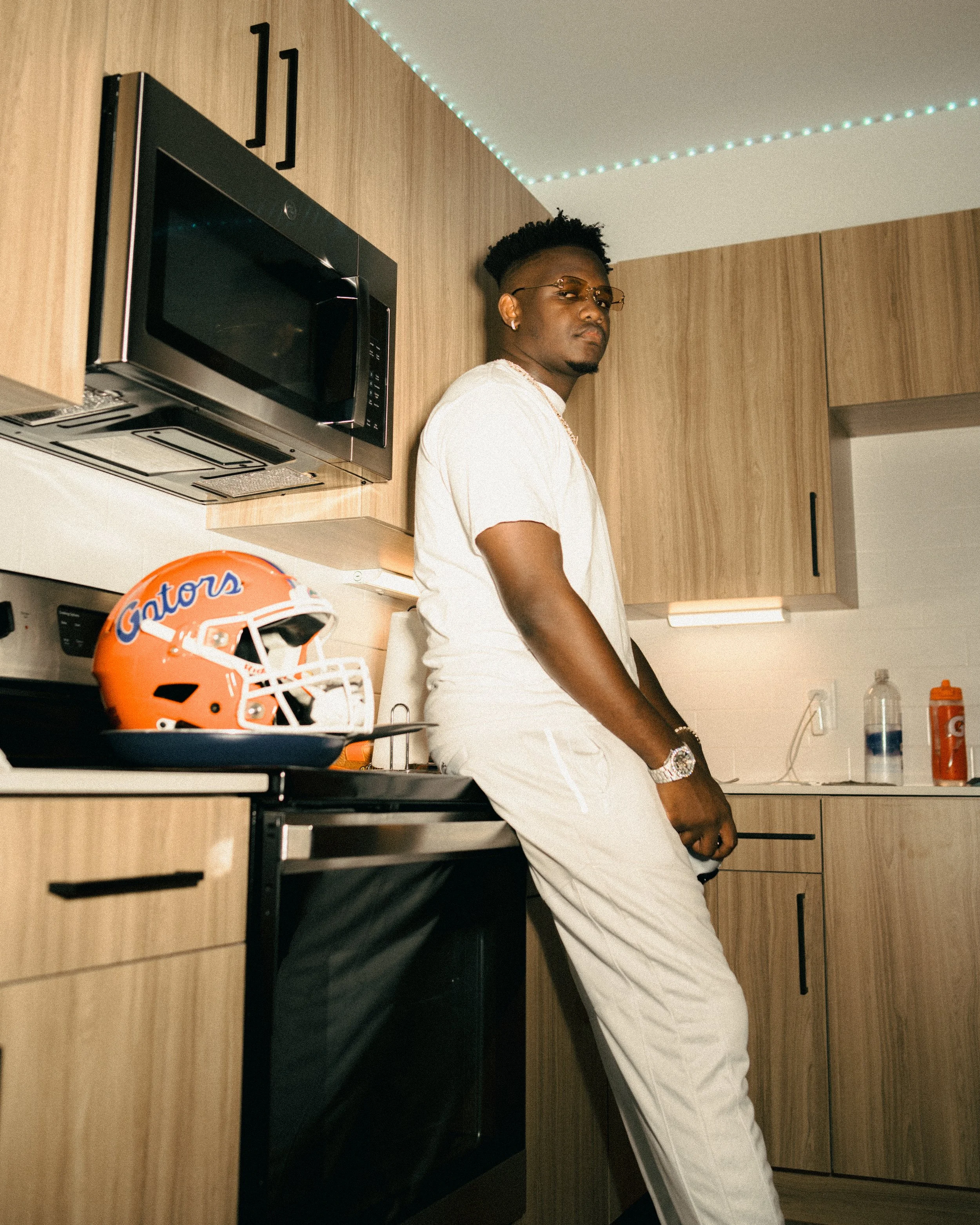 Young man in white athletic clothes posing in a modern kitchen with a Florida Gators football helmet on the counter.