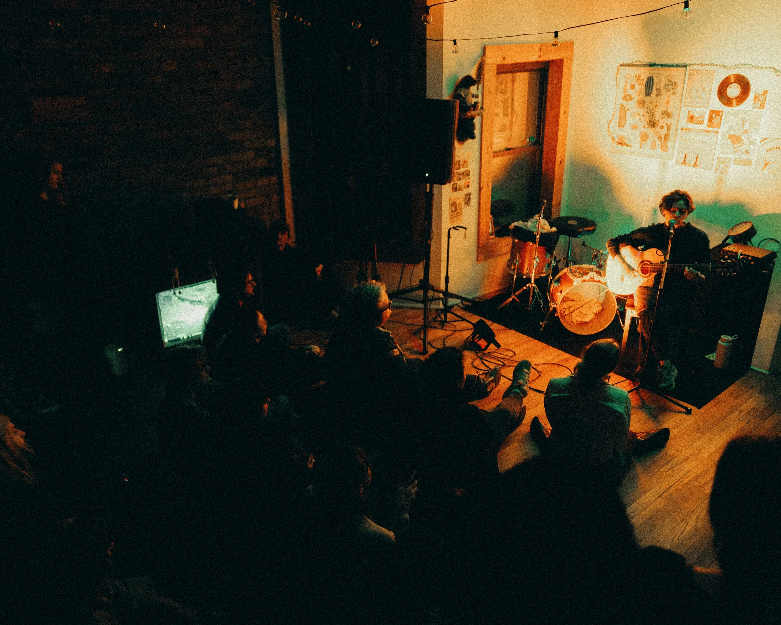 A musician performs with an acoustic guitar on a small stage in a cozy venue, illuminated by warm lighting, with an audience seated on the floor.