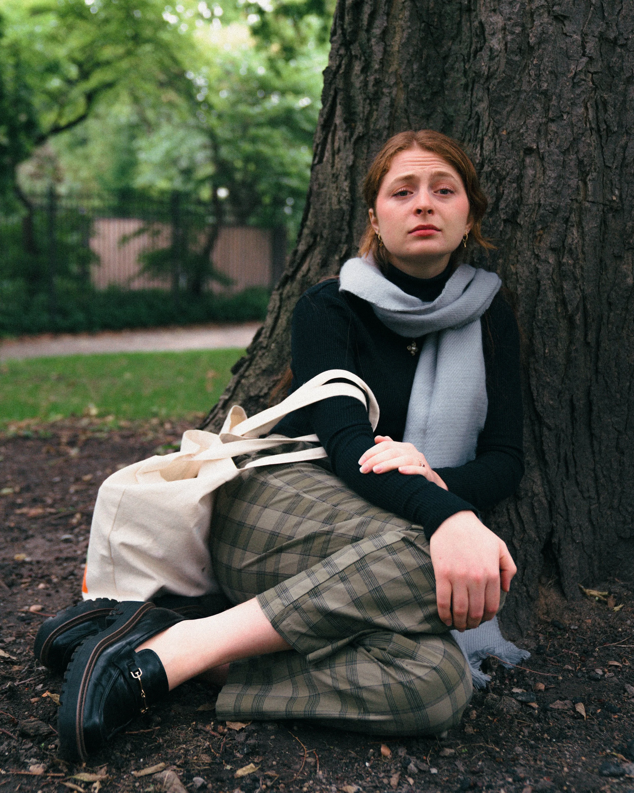 A young woman with red hair sitting on the ground and leaning against a large tree trunk in a park, with a somber expression.