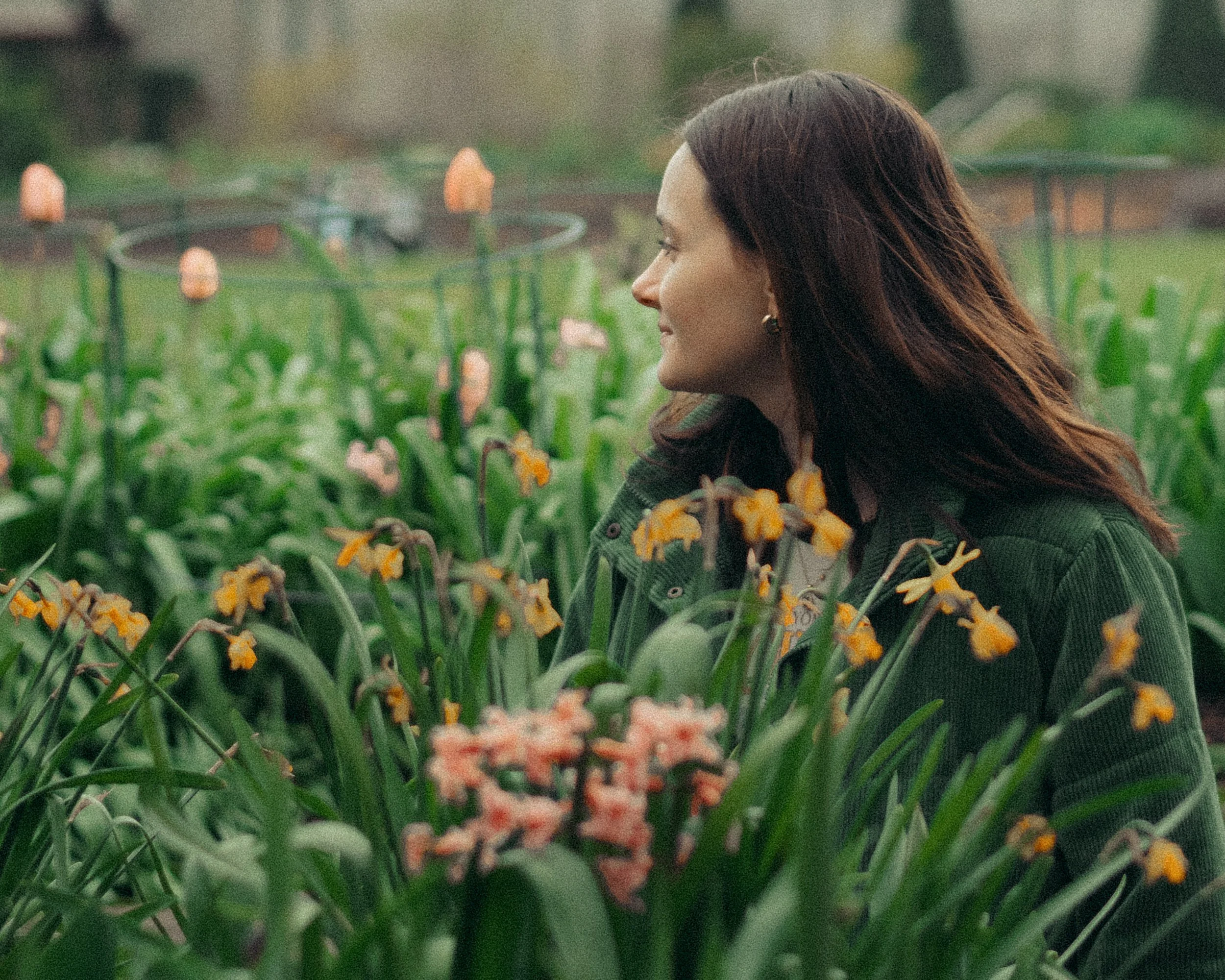 A woman with long dark hair wearing a green jacket standing among orange and pink flowers in a garden.
