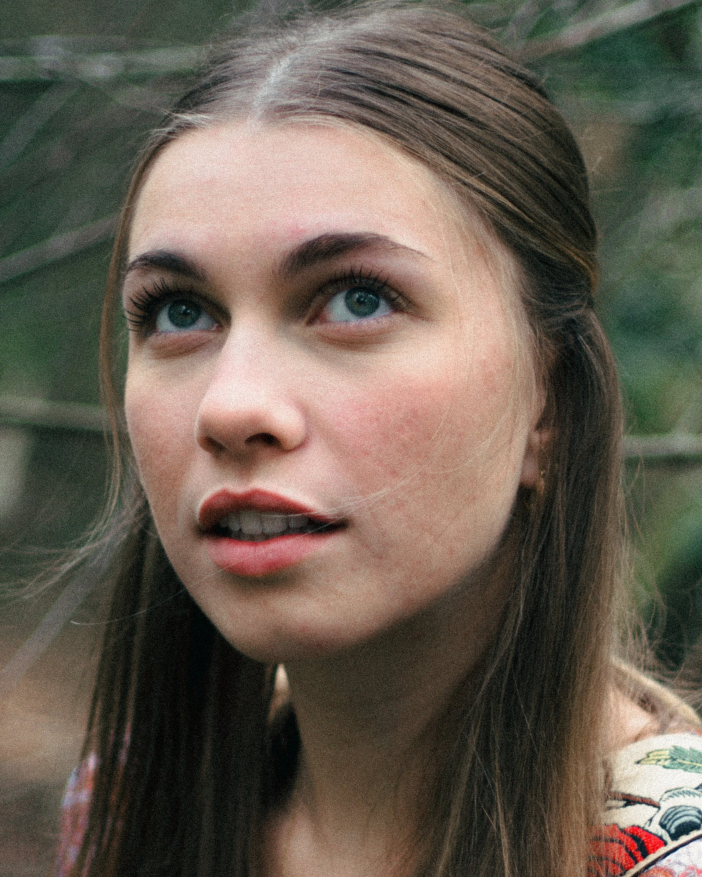 Close-up of a young woman with blue eyes and brown hair, outdoors, surrounded by green foliage.