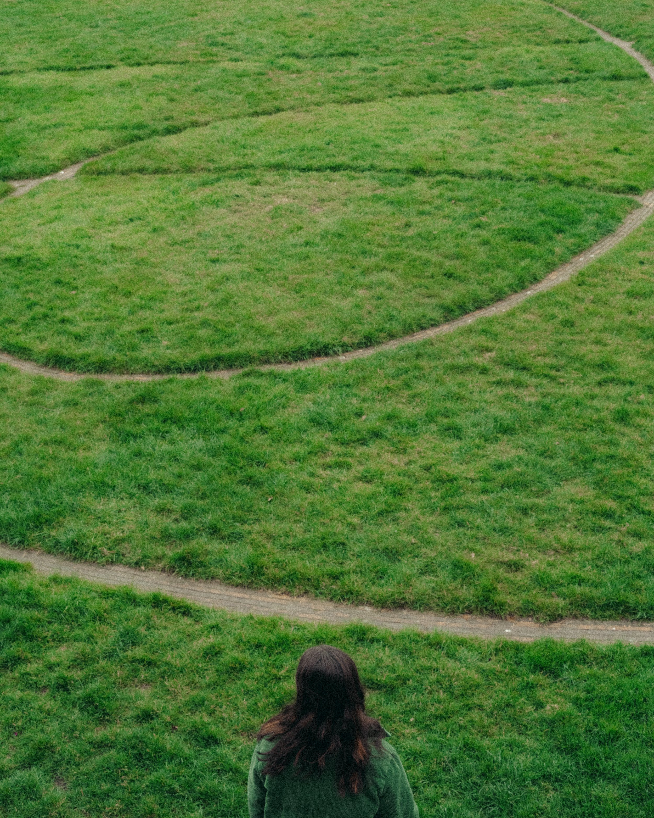 A woman with long dark hair, wearing a green jacket, stands on a grassy hill overlooking a park with winding paths and green lawns.