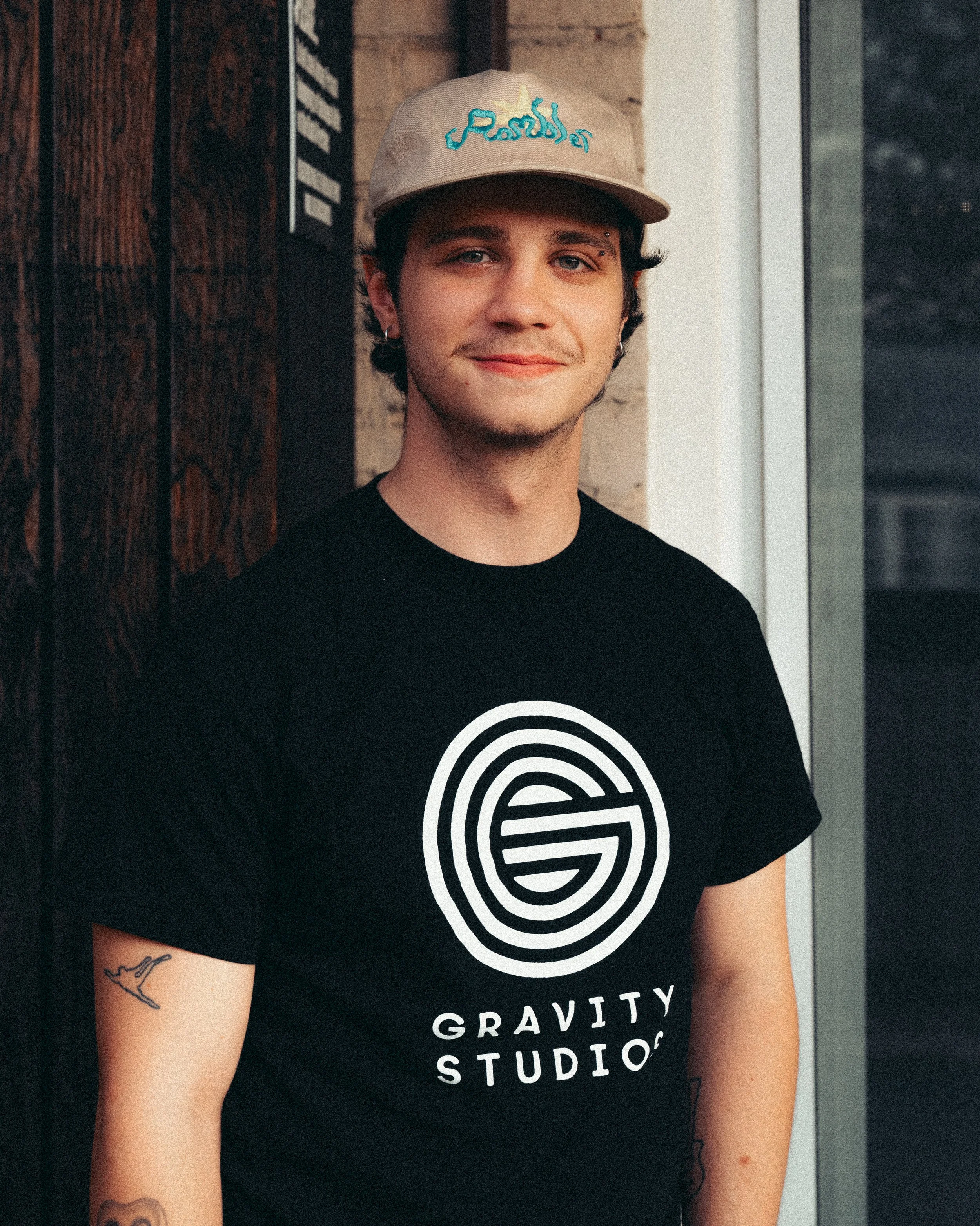 A young man with earrings and tattoos, wearing a beige baseball cap with blue writing and a black T-shirt with a white circular logo and the words "Gravity Studio," standing outdoors against a brick wall with a window nearby.