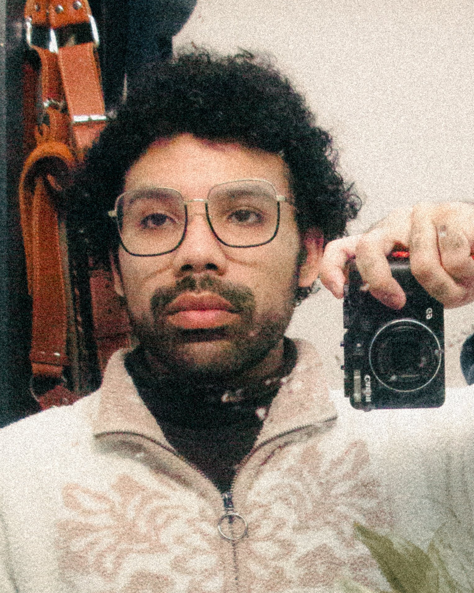 A man with curly hair, glasses, and a mustache is taking a mirror selfie with a camera. He is wearing a beige sweater with a butterfly design and a black shirt underneath. There is a wooden object or piece of furniture in the background.