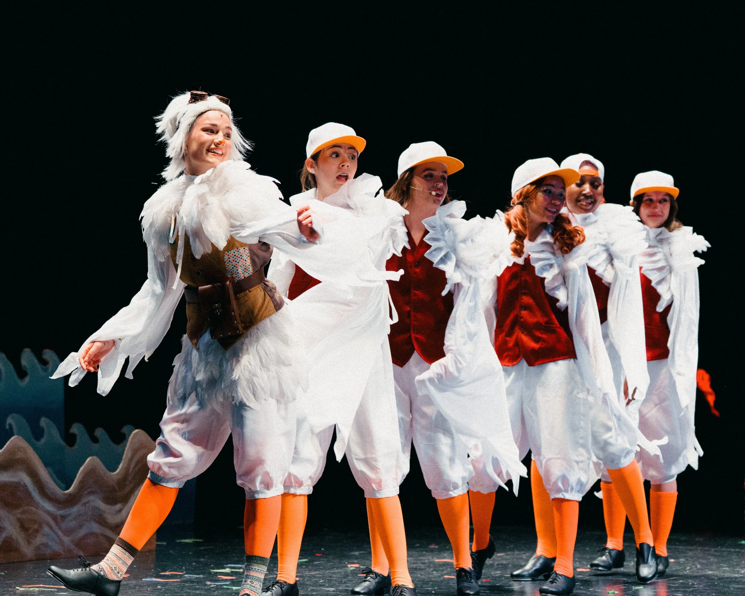 Theatrical performance featuring six women dressed as birds with feathers and orange beak-like hats, standing on stage against a dark backdrop with a wavy set piece.