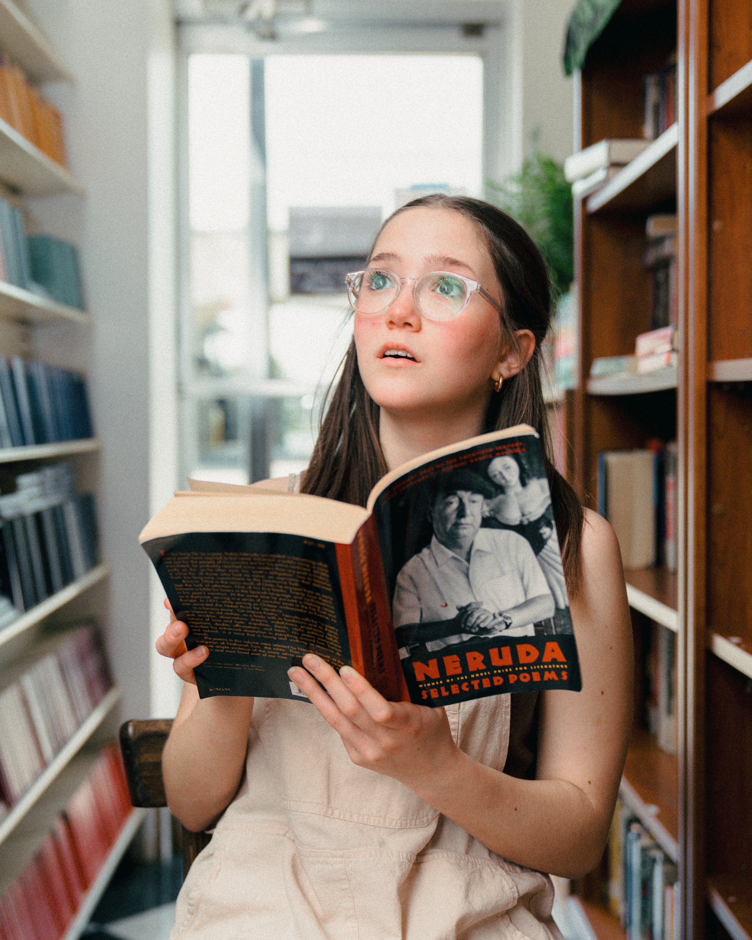 A young woman with glasses reading a book titled "Neruda" in a library, sitting between bookshelves filled with books.