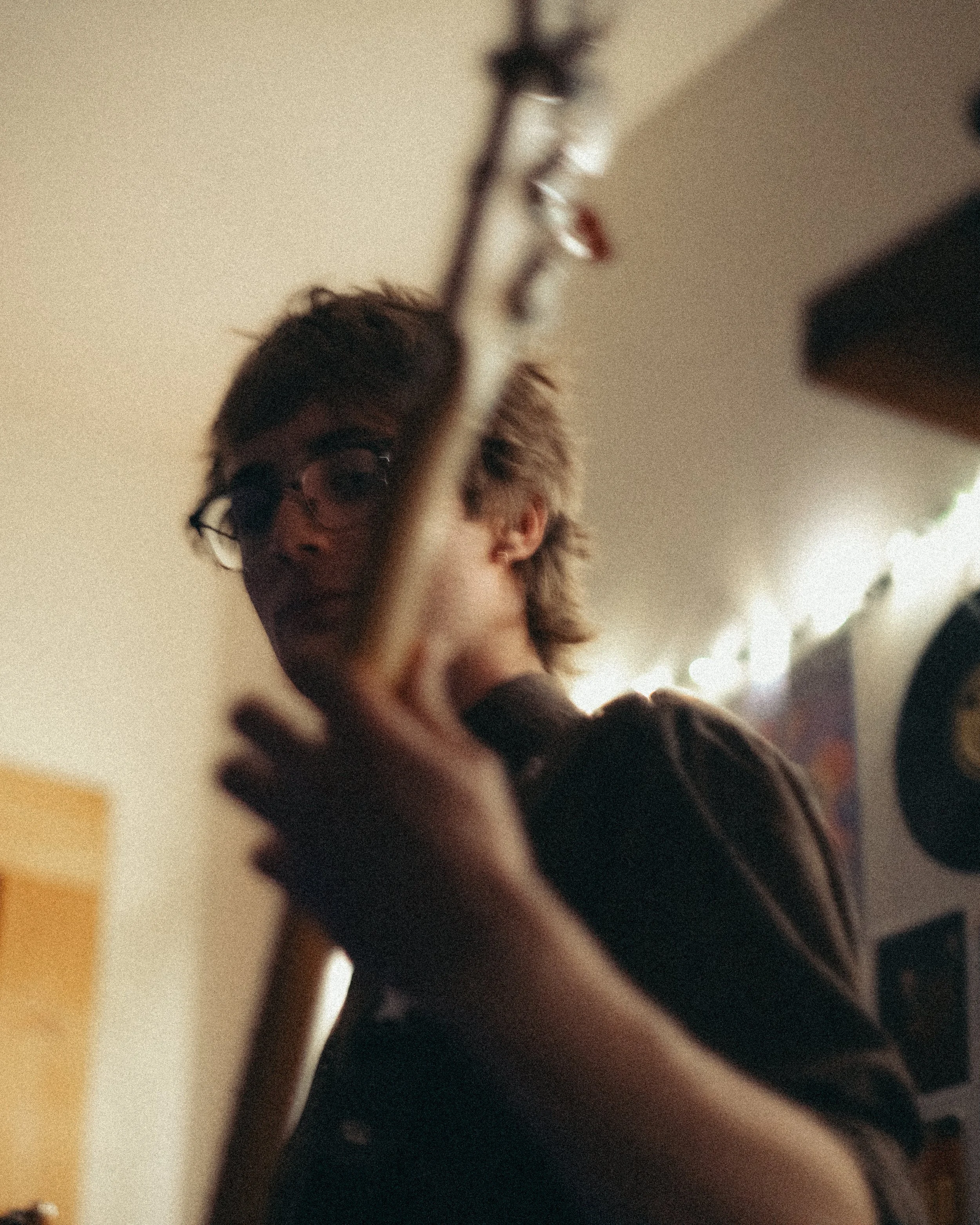 A person with glasses and medium-length hair is seen in a dimly lit room, with their face partially obscured, holding an object with their hand, with background lights and decor.