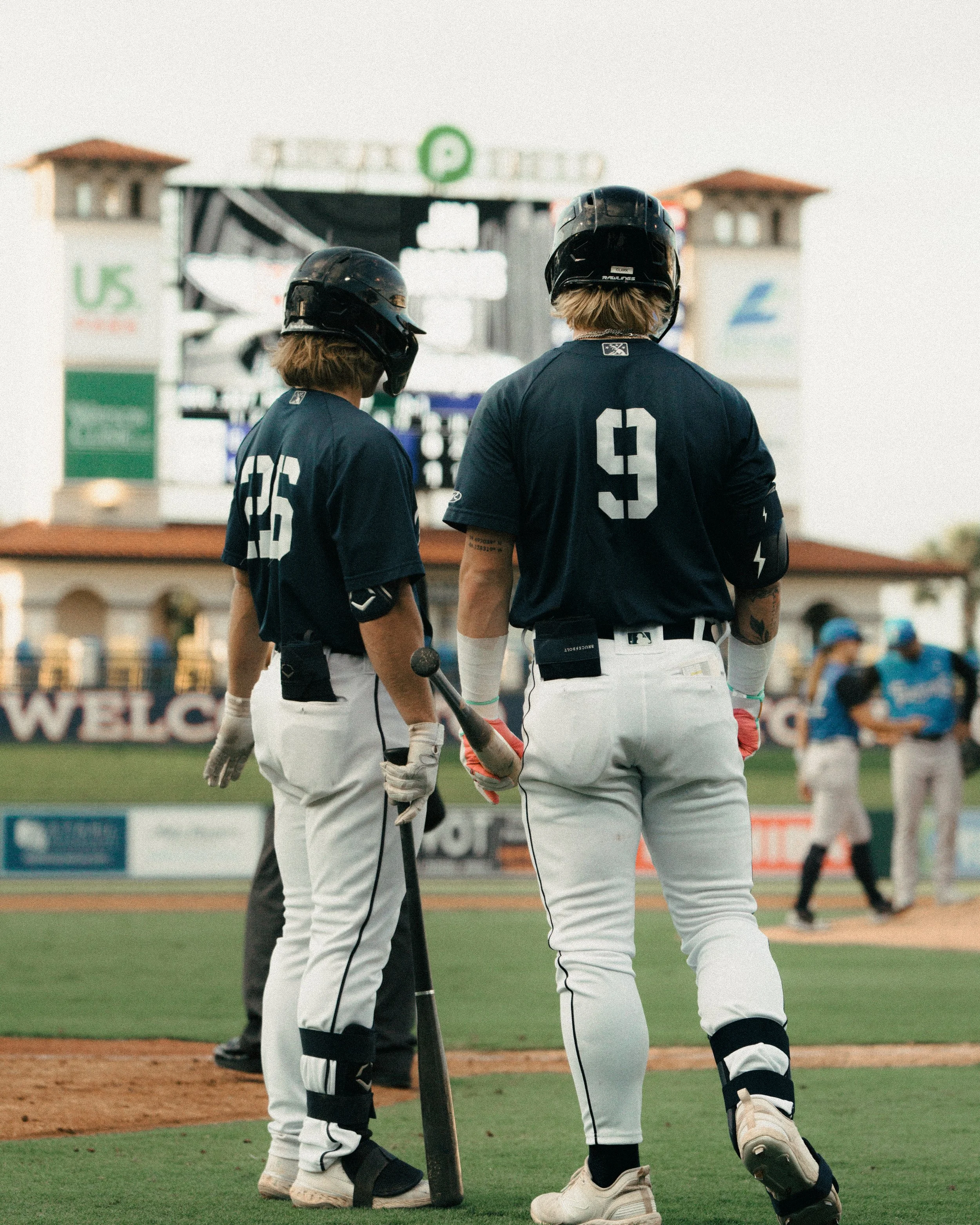 Two baseball players in navy jerseys and white pants talking on the field, with a scoreboard and other players in the background.