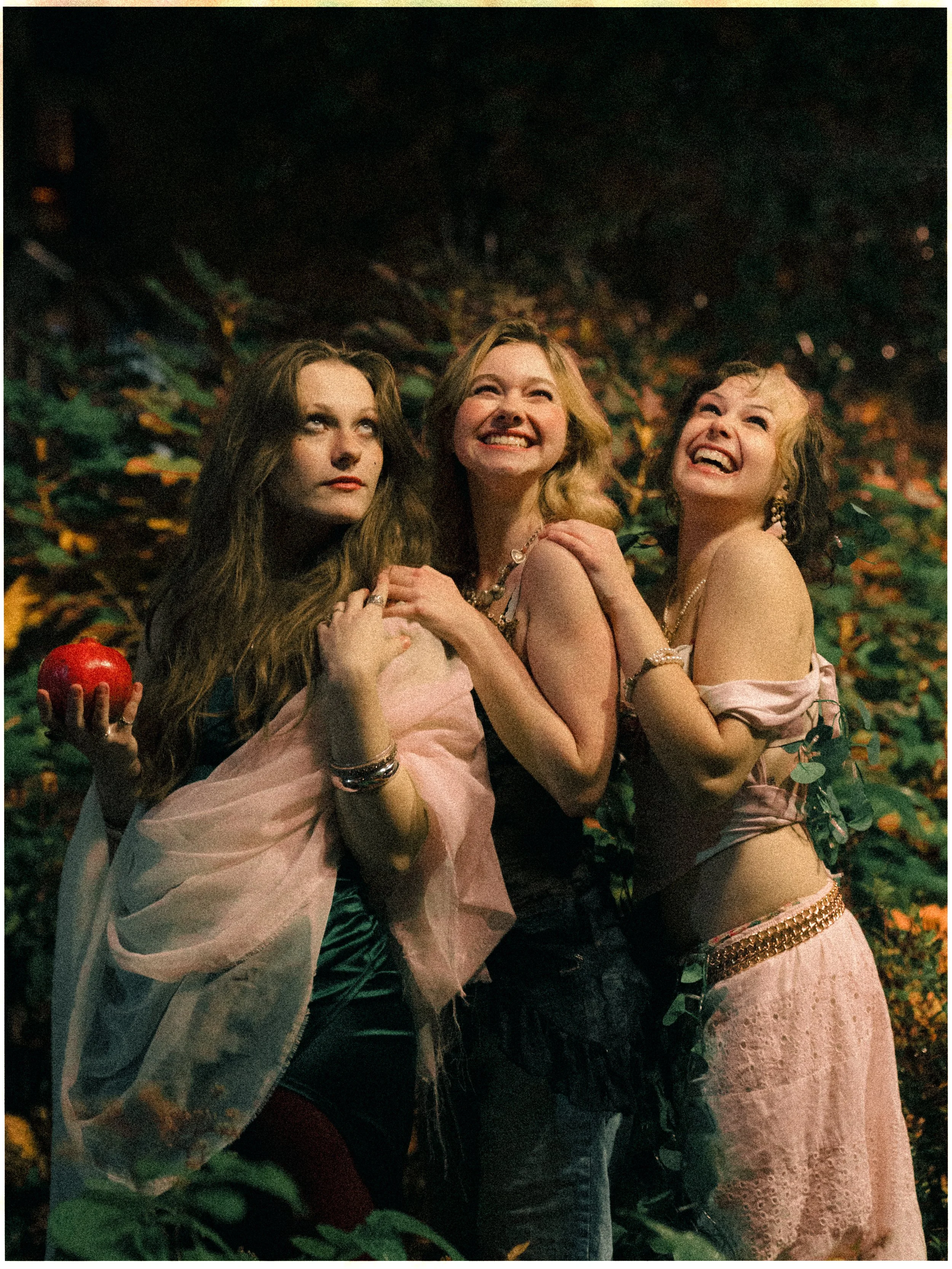 Three women standing close together outdoors at night surrounded by foliage, with two of them smiling and one looking serious, the woman on the left holding a red apple.