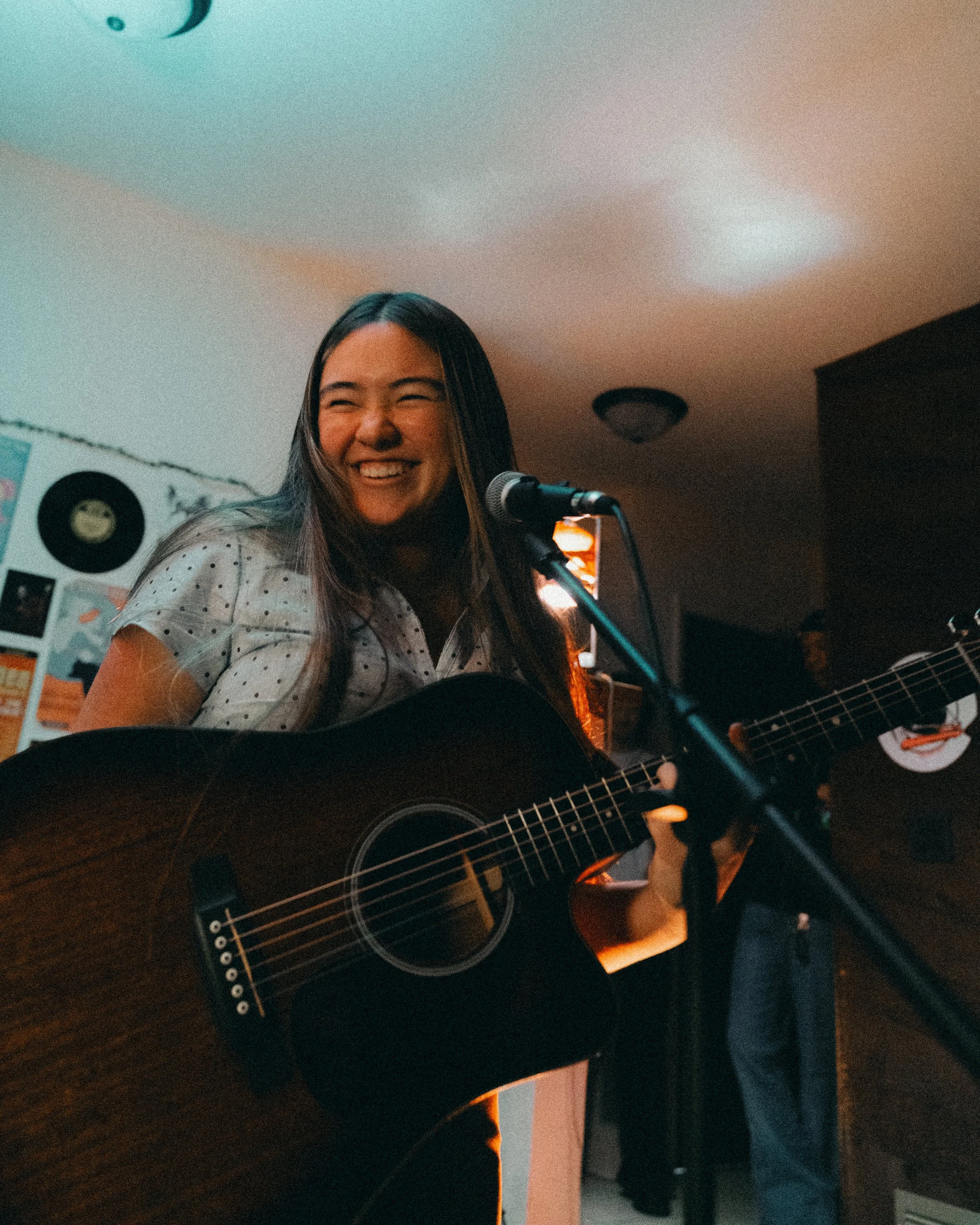 A woman smiling and playing an acoustic guitar in a room decorated with vinyl records and posters.