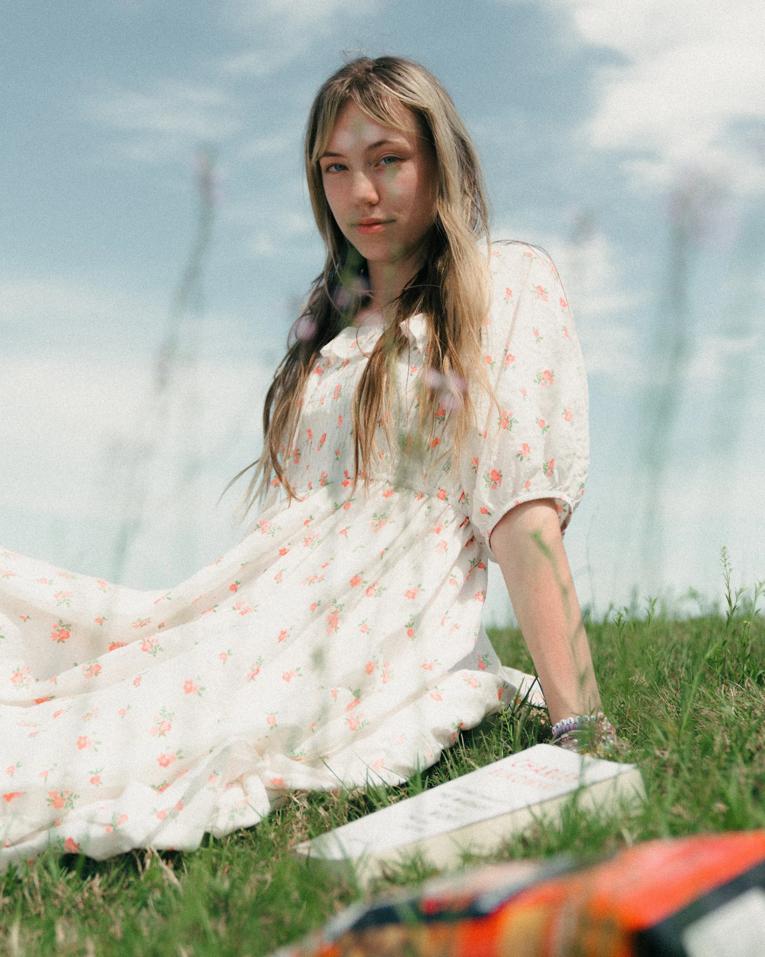 A young woman sitting on grass outdoors in a white dress with pink flowers, looking at the camera with a slight smile, with blue sky and clouds in the background.