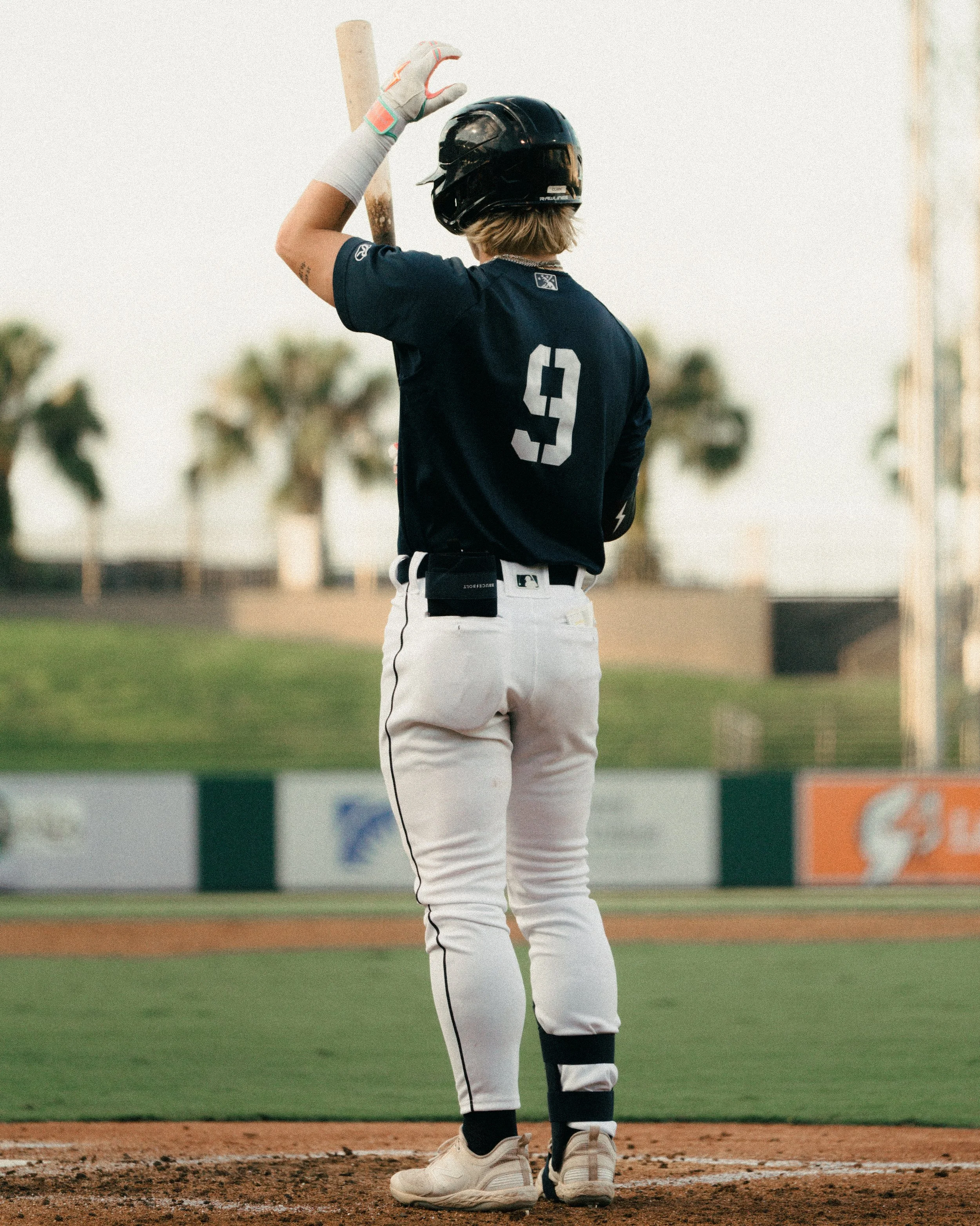 Baseball player standing on home plate with bat, wearing helmet, navy jersey with number 9, white pants with black stripe, and black athletic tape on ankle.