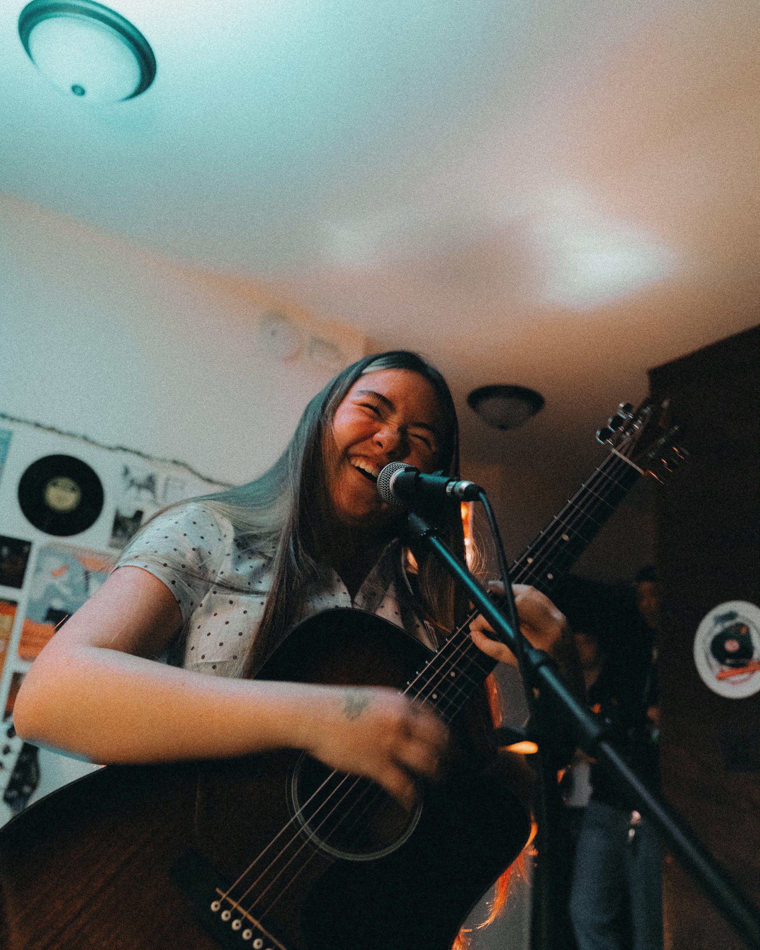 A woman with long dark hair singing and playing an acoustic guitar in a cozy indoor setting, with vinyl records on the wall behind her.