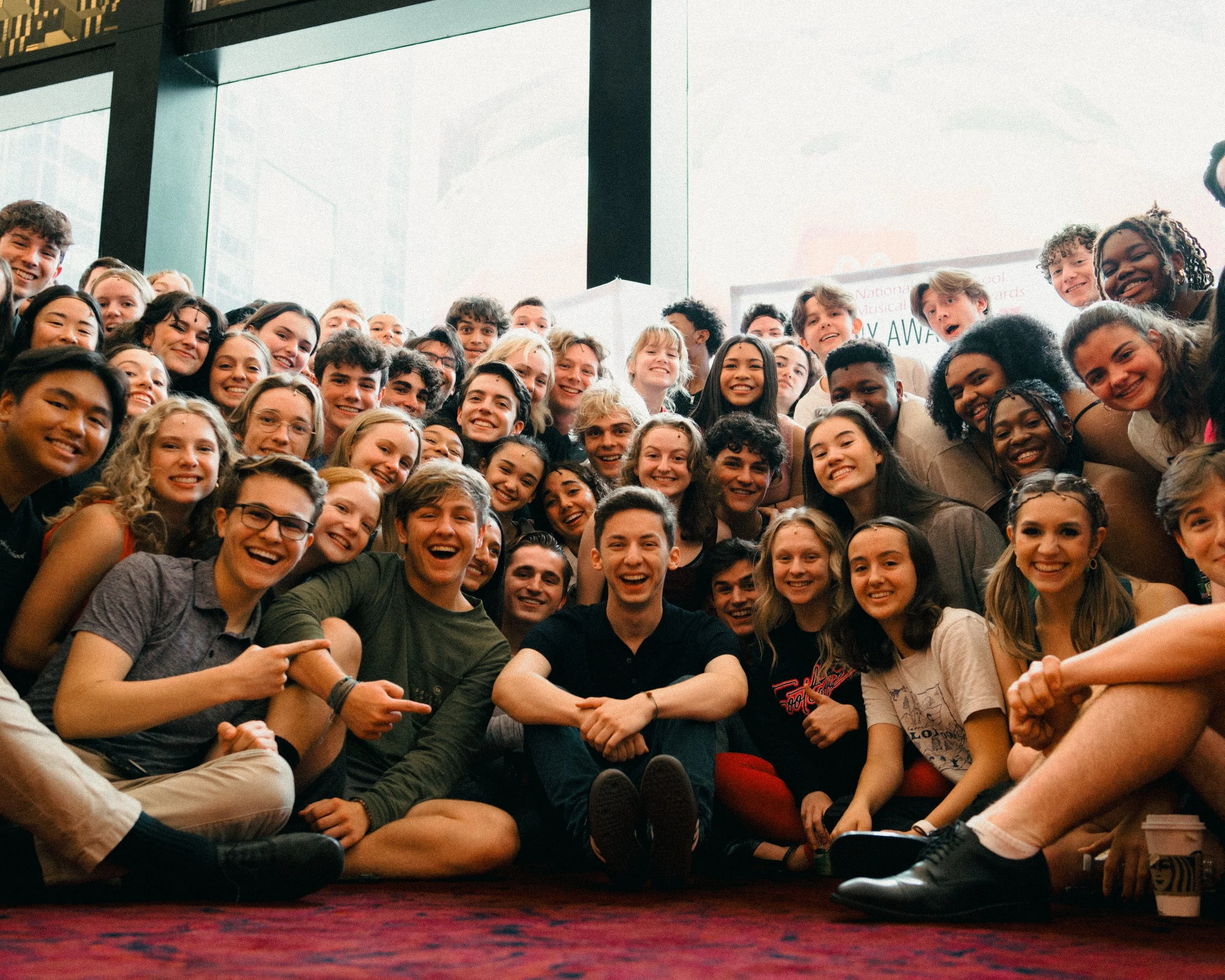 Large group of smiling young people gathered together indoors, sitting on a carpeted floor, with a large window behind them.