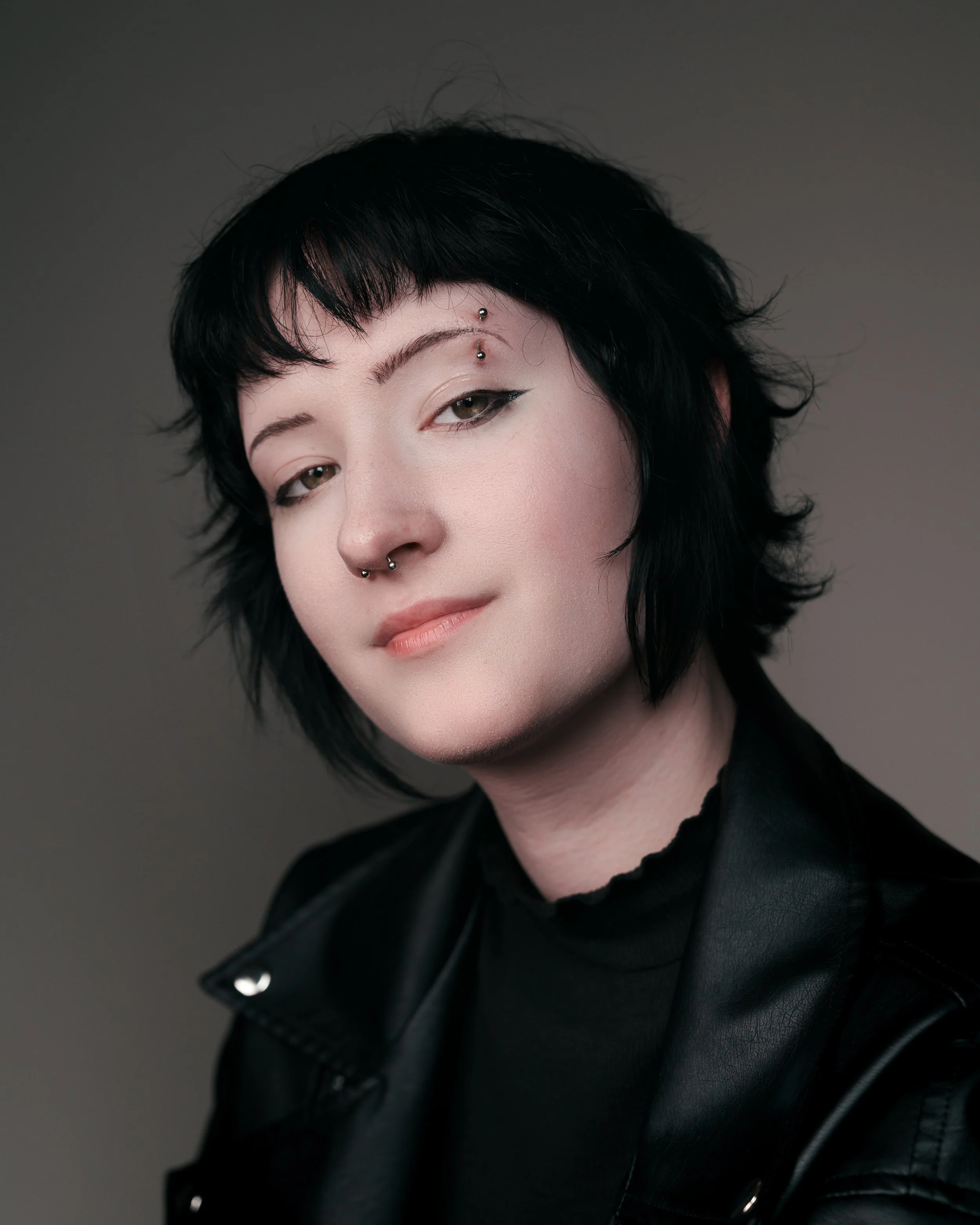 Portrait of a woman with black hair and piercings, wearing a black leather jacket and black top, looking confidently at the camera.