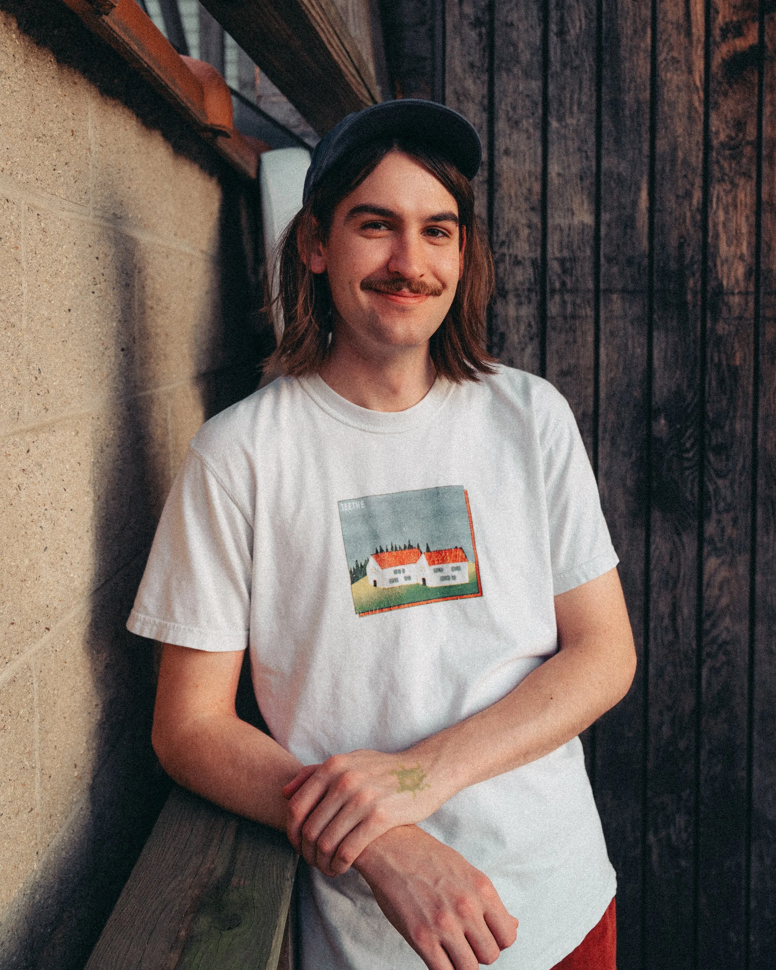 A young man with shoulder-length brown hair, mustache, and wearing a black cap, white T-shirt with a house illustration, and red pants, standing outdoors against a wooden wall and beige concrete wall.