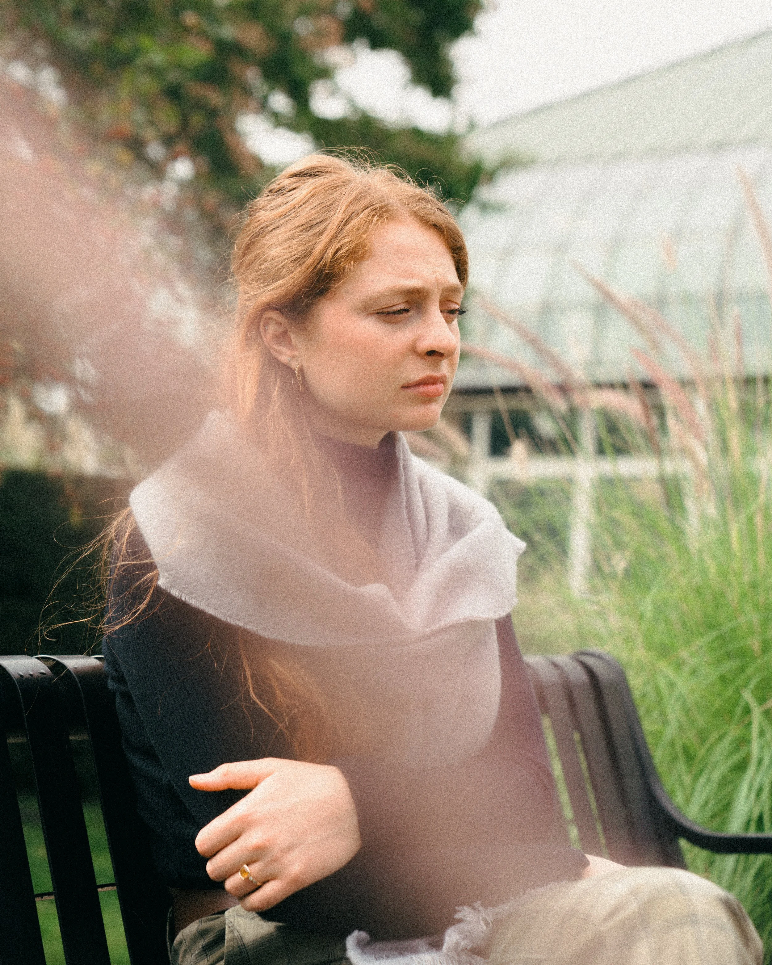 A young woman with red hair sitting on a bench outdoors, looking sad or contemplative, with greenery and a greenhouse in the background.