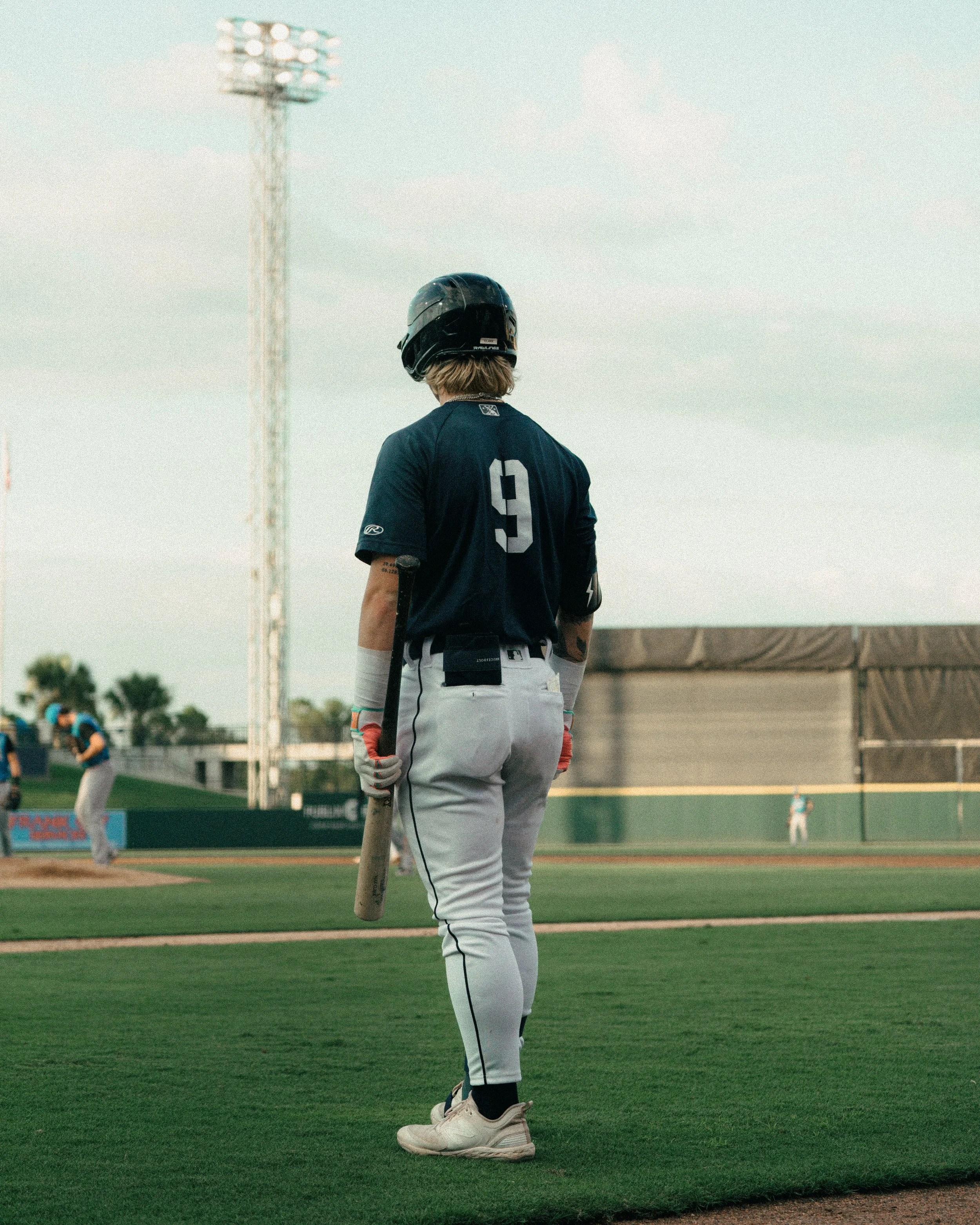 A baseball player wearing a black helmet, navy blue jersey with the number 9 on the back, gray pants, and batting gloves stands holding a bat, looking towards the field during a game.