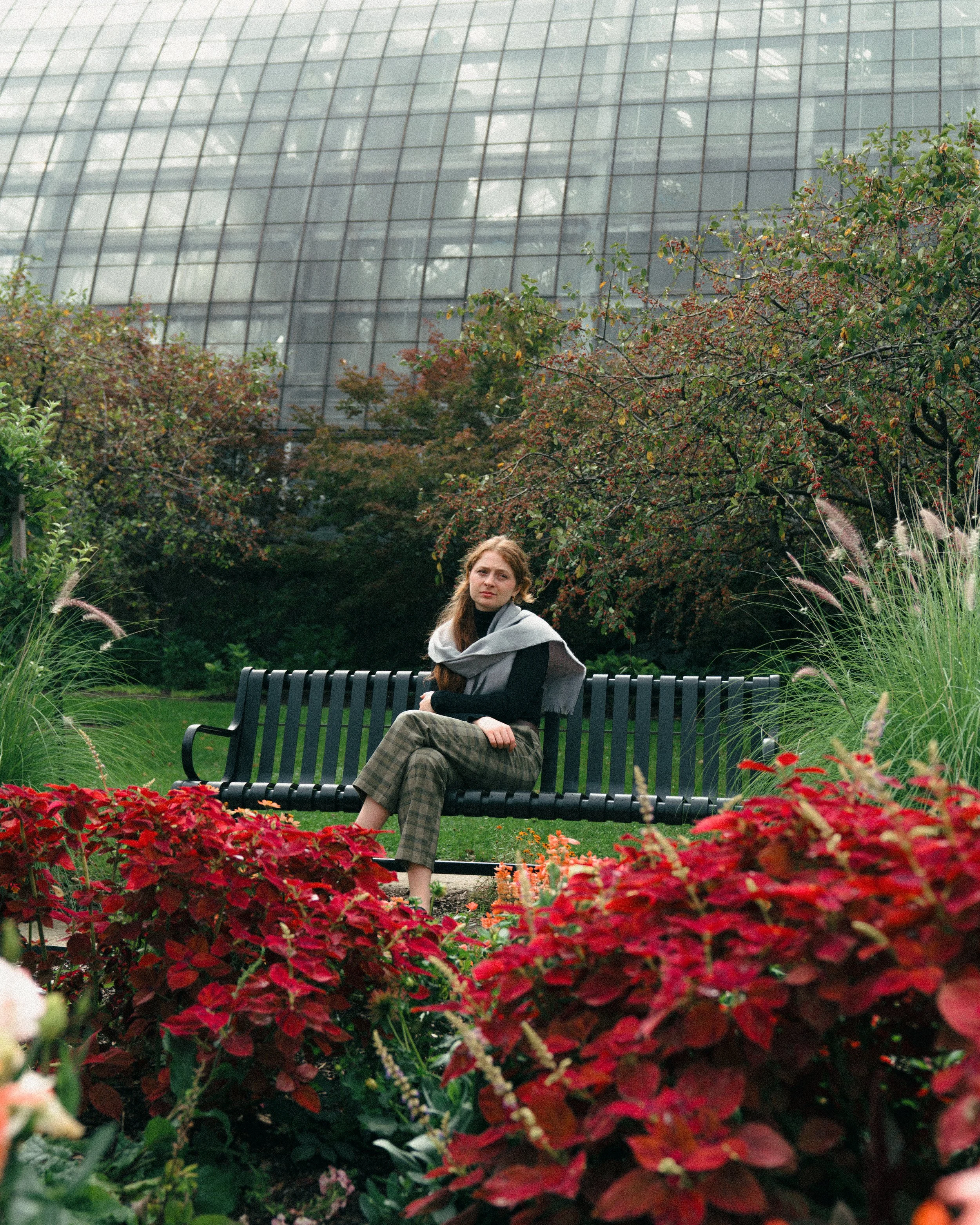 A woman sitting on a park bench surrounded by colorful flowers and plants, with a modern glass building in the background.