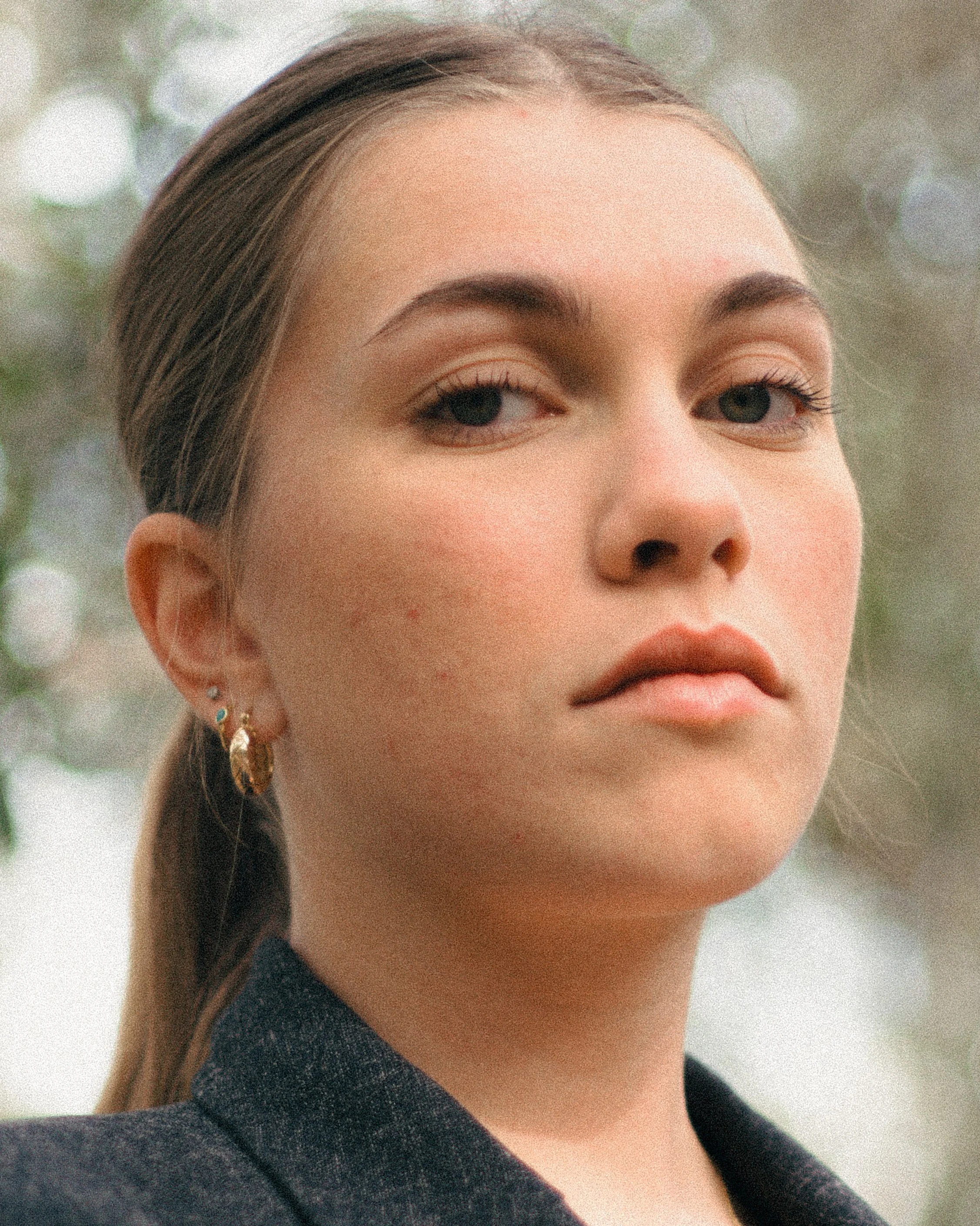 Close-up of a young woman with brown hair, wearing earrings and a dark blazer, outdoors with blurred background.